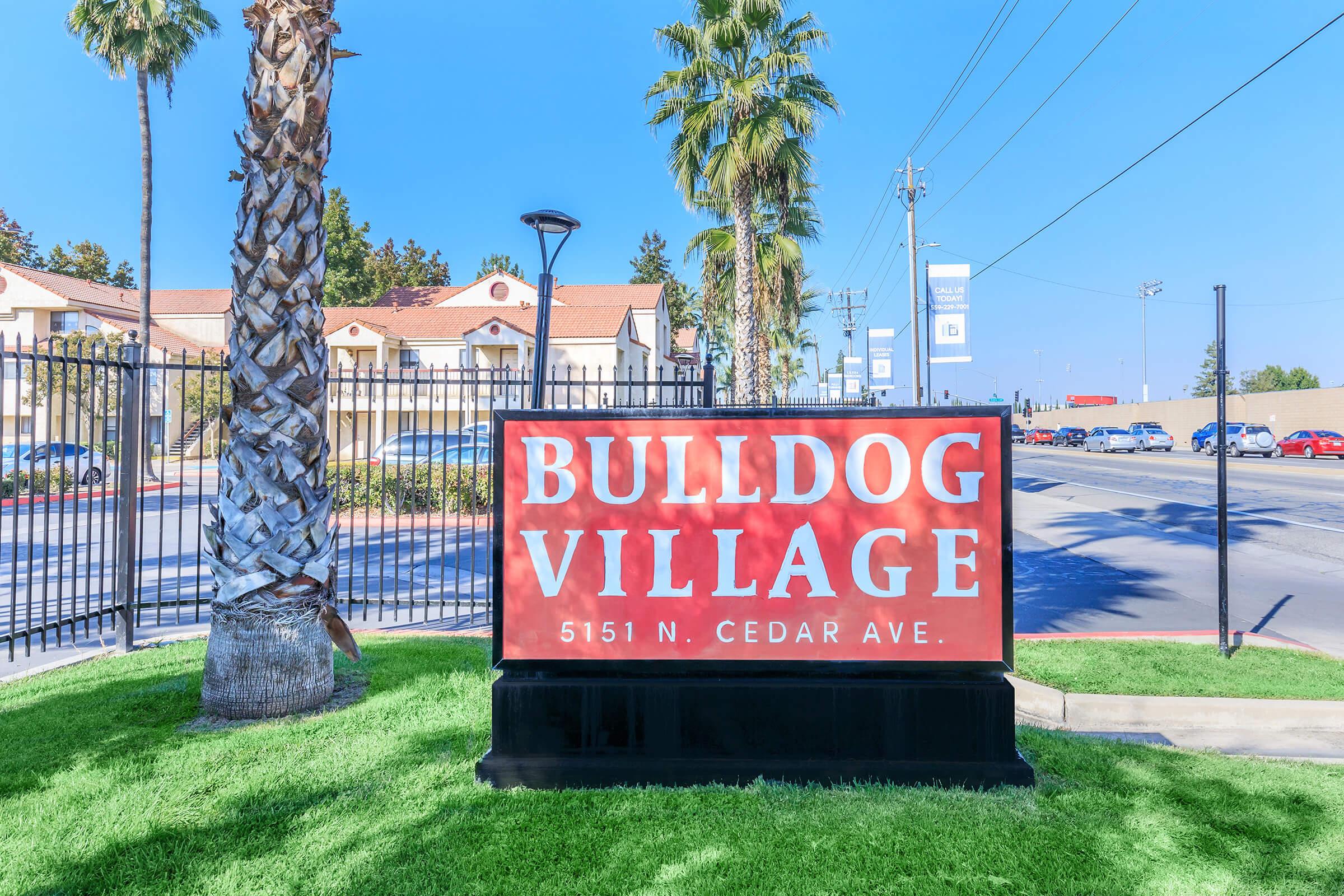 Sign for Bulldog Village located at 5151 N. Cedar Ave, featuring a red background with large white text. Surrounding the sign are palm trees and green grass, with a clear blue sky. The area reflects a sunny, vibrant atmosphere.