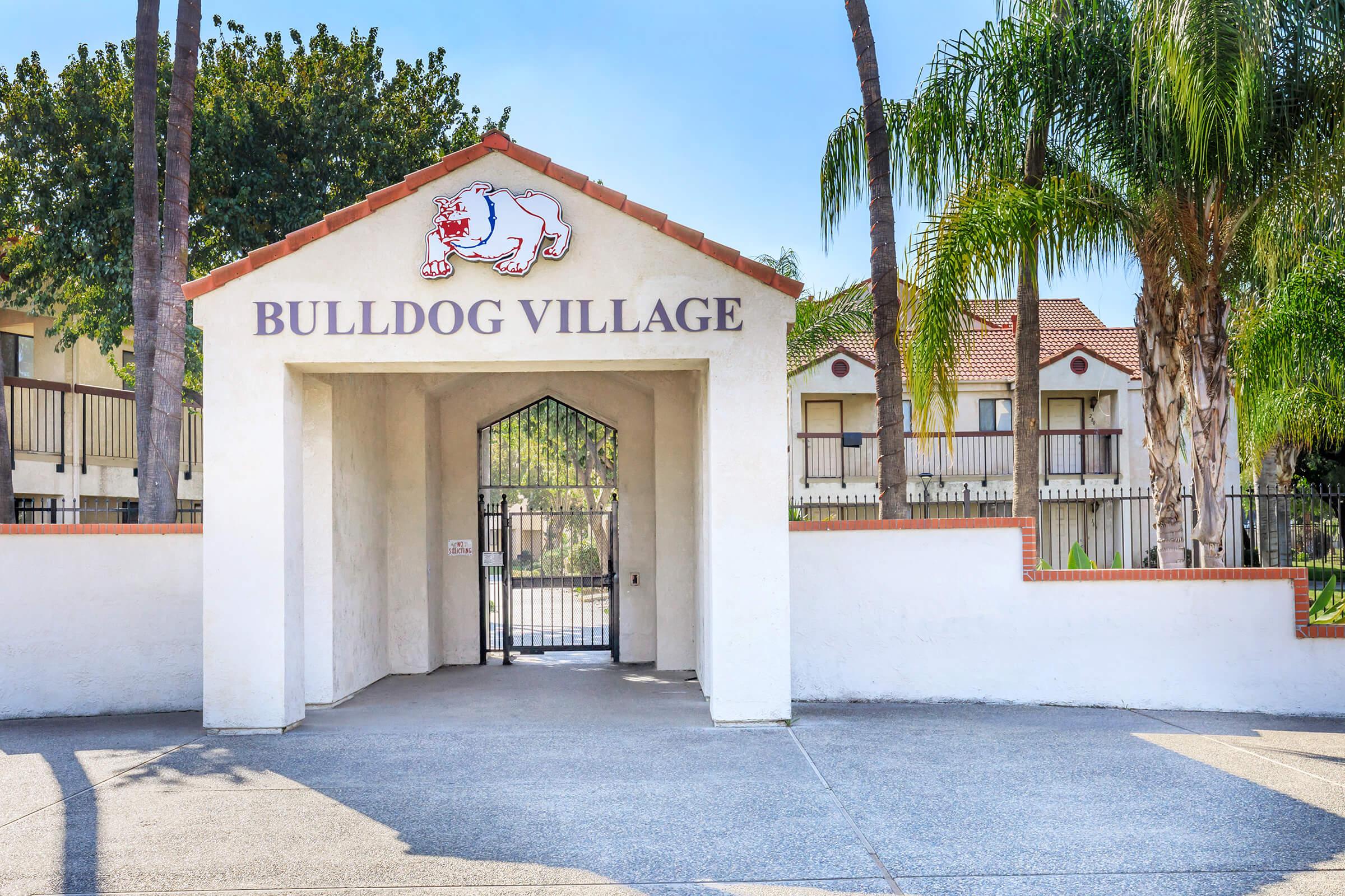 Entrance to Bulldog Village, featuring a sign with a bulldog logo. The structure is painted white with a red-tiled roof and surrounded by palm trees. A paved pathway leads through the archway, and a gated entrance is visible. The background includes buildings typical of residential complexes.
