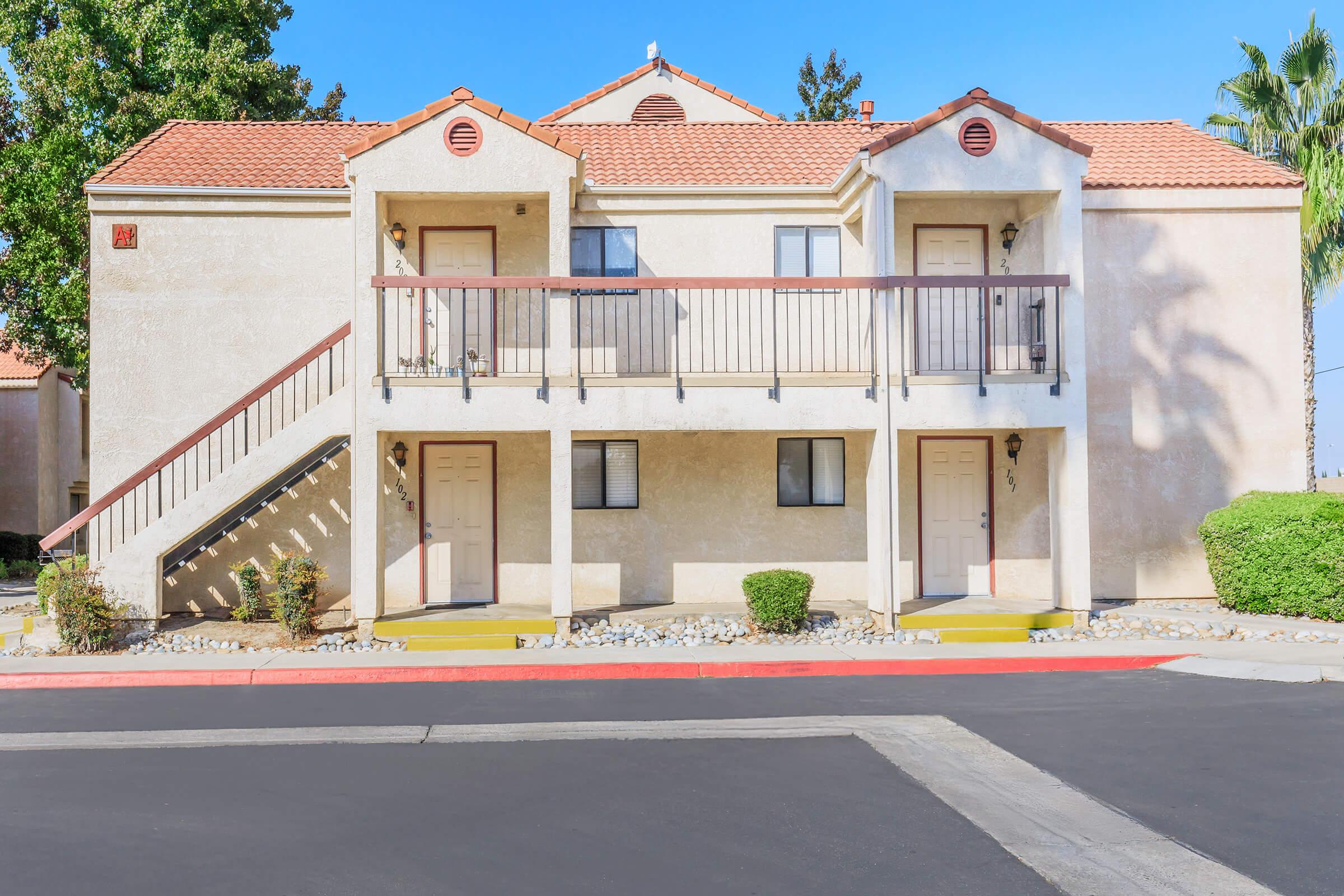 A two-story apartment building with a beige exterior, featuring a staircase leading to the upper units. Each unit has a balcony and two doors on the ground floor. The surrounding area includes shrubs and palm trees, with a clear blue sky above. The pavement in front is marked.