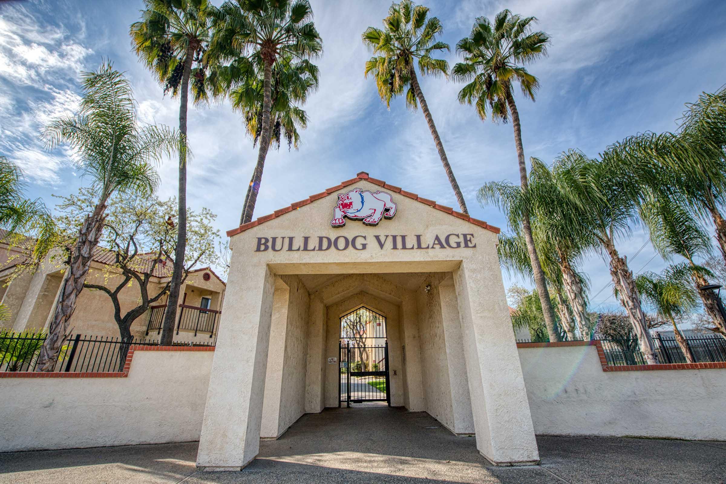 Entrance to Bulldog Village featuring a decorative archway with "BULLDOG VILLAGE" displayed prominently above. The area is surrounded by palm trees and other greenery, under a bright blue sky with some clouds. The overall scene is inviting and represents a campus or community setting.