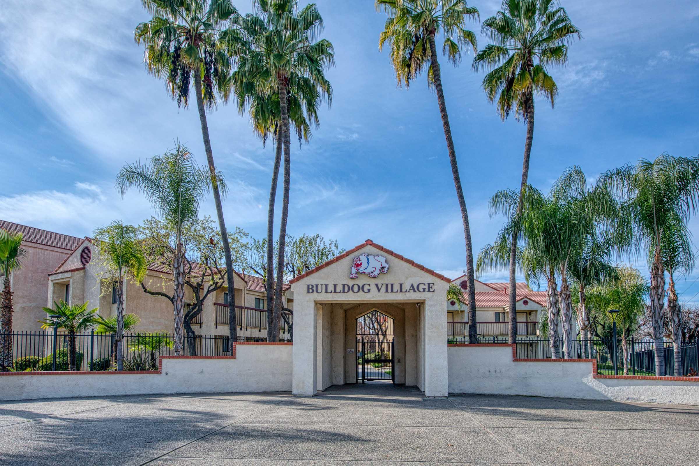 Entrance to Bulldog Village, framed by palm trees and lush greenery. The archway features the name "BULLDOG VILLAGE" prominently displayed. The setting is bright and inviting, with a clear blue sky above and well-maintained surroundings, suggesting a residential or community area.