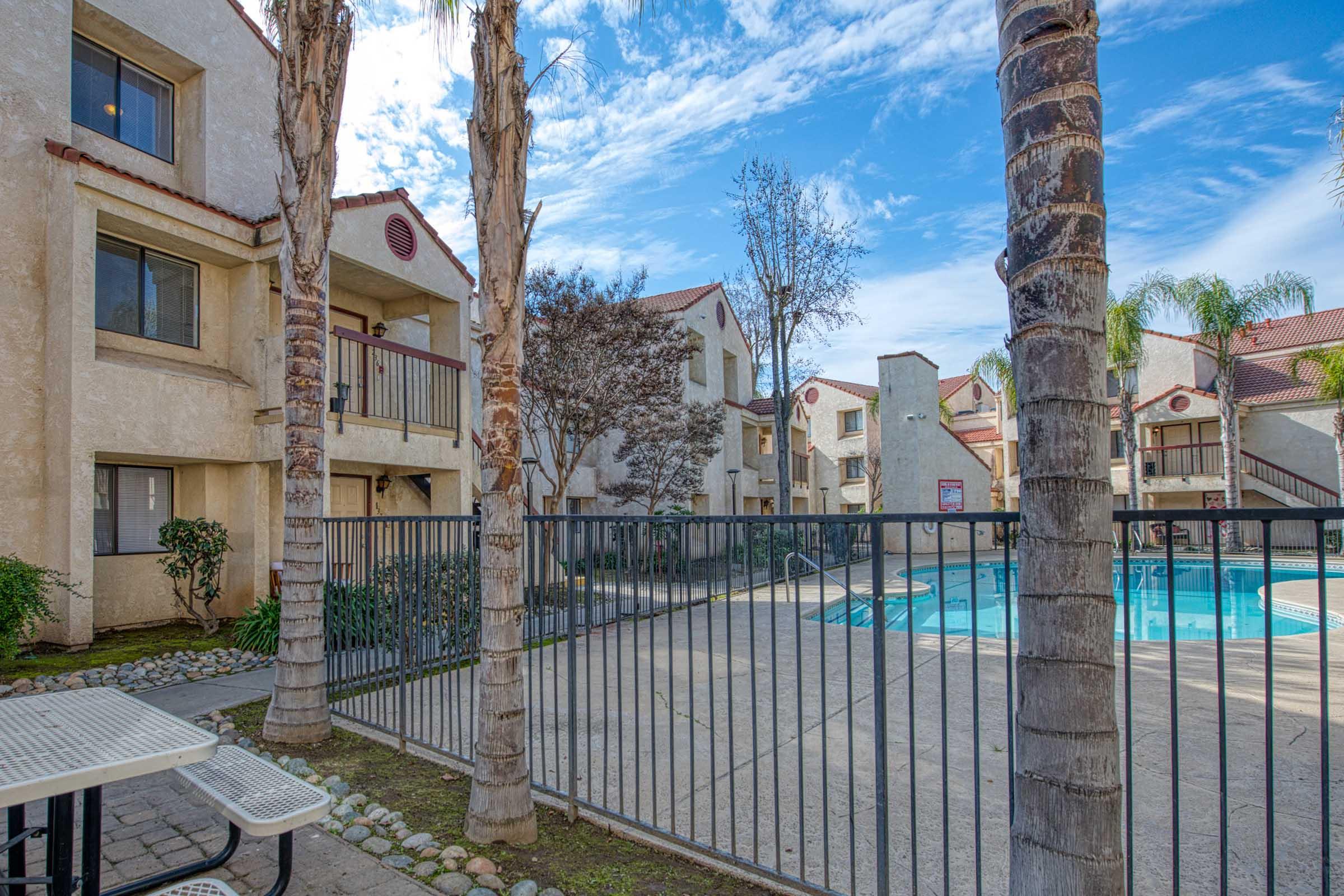 View of an apartment complex with three-story buildings surrounding a swimming pool. Palm trees and shrubbery are visible in the foreground, and the sky is clear with a few clouds. A fenced area includes picnic tables nearby, creating a relaxed outdoor space for residents.