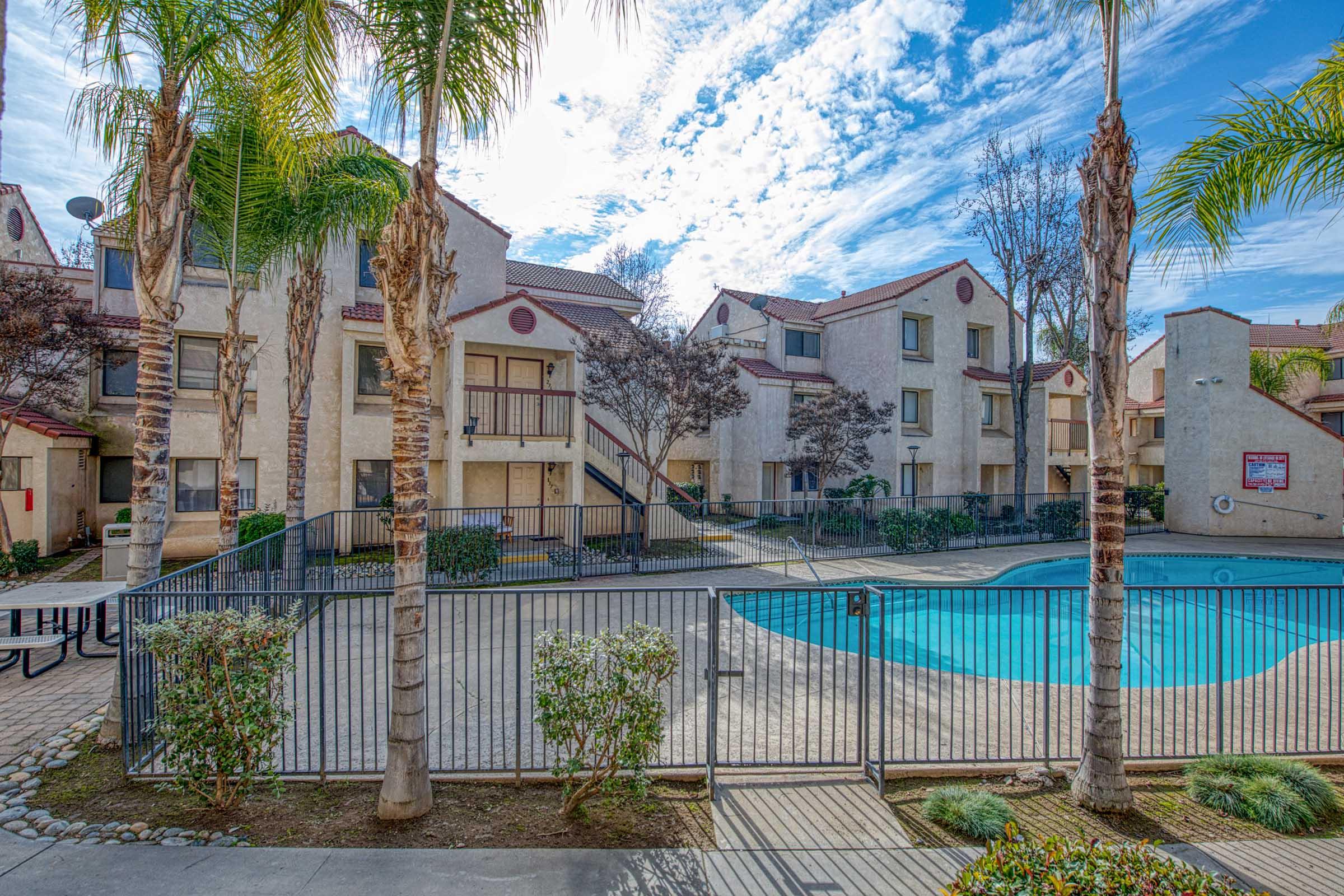 A view of a residential apartment complex featuring a swimming pool surrounded by a fenced area. Palm trees and shrubs are visible in the foreground, with a two-story building in the background, under a partly cloudy blue sky.