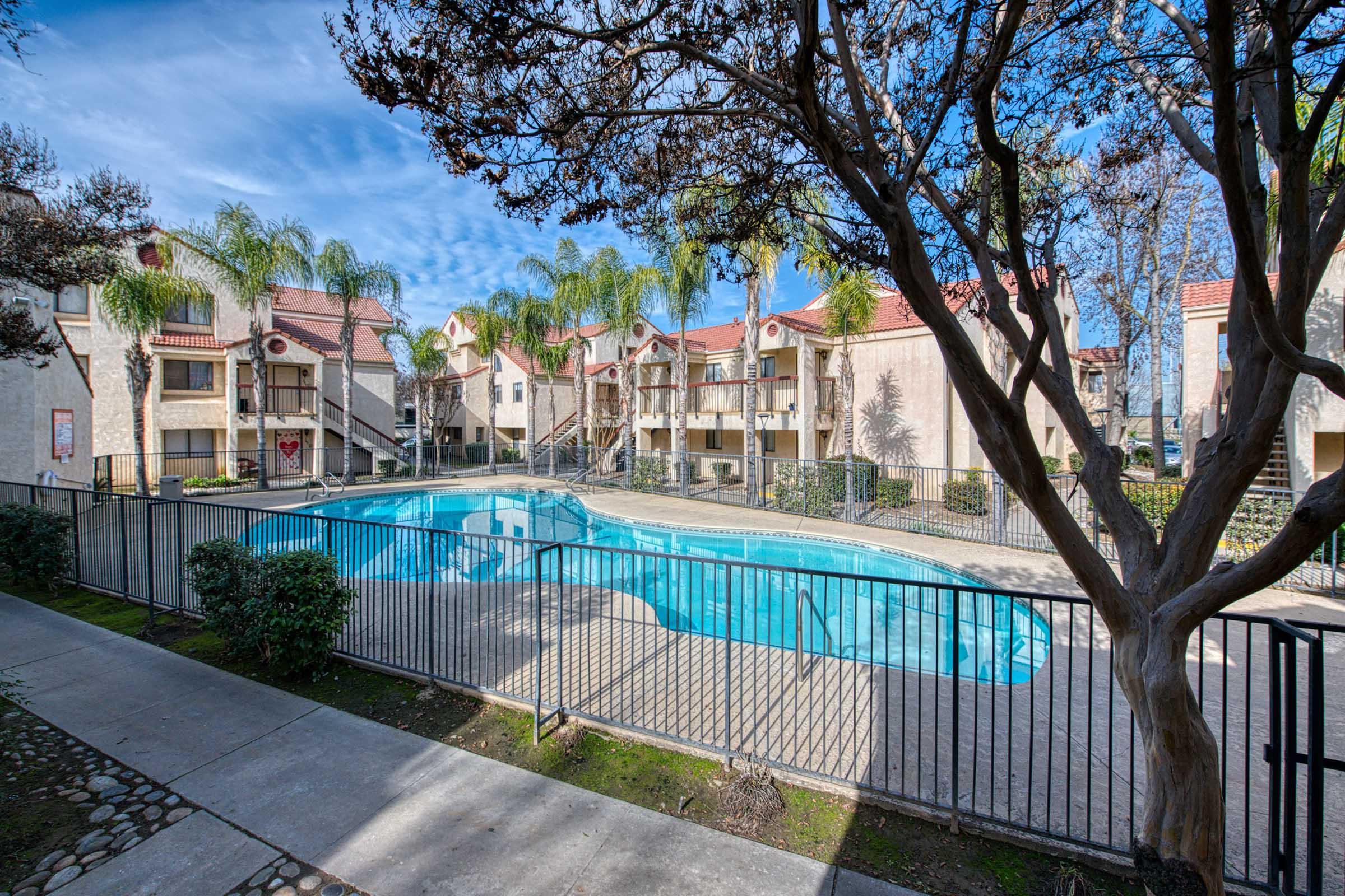 A clear view of a residential apartment complex featuring an inviting swimming pool surrounded by palm trees. The area is enclosed by a black fence, and several apartment buildings can be seen in the background under a bright blue sky with scattered clouds.