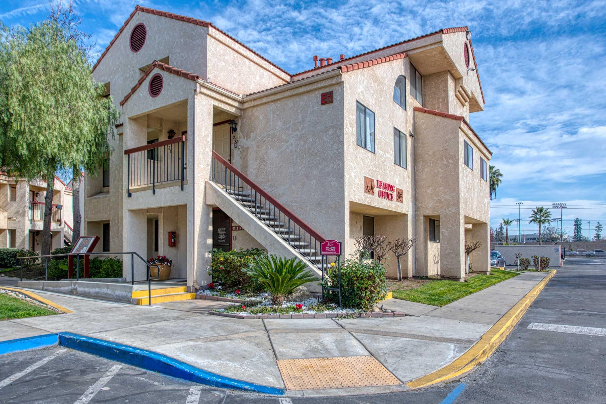 A two-story apartment building with a light-colored exterior, red accents, and a staircase leading to the second floor. Surrounding greenery and a parking lot are visible, under a partly cloudy blue sky. A sign indicates the building is associated with a leasing office.