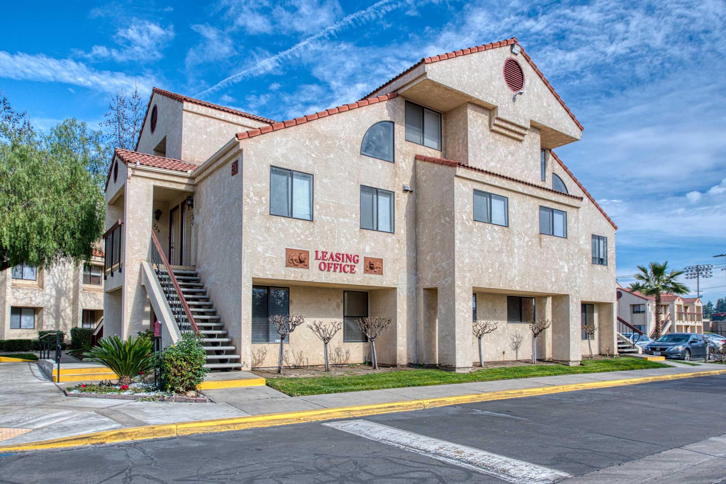 A multi-story building with a beige exterior featuring a leasing office sign in red. There are two staircases leading to the upper levels, small trees and plants landscaping the front. The sky is partly cloudy, adding a bright atmosphere to the scene.