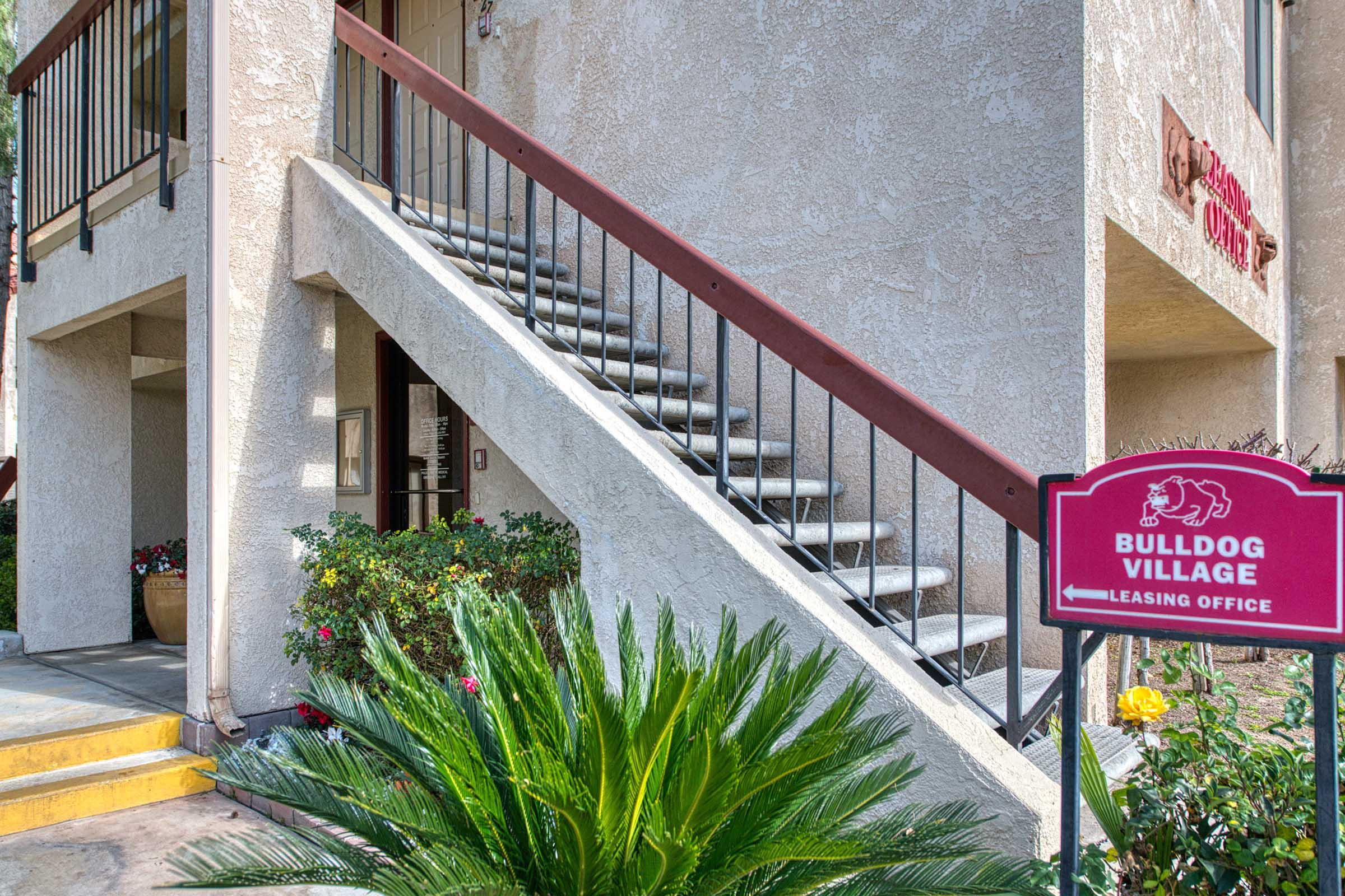 Stairs leading to a second-floor entrance with a railing, next to a sign reading "Bulldog Village Leasing Office." The area features a plant in the foreground and colorful flowers along the path. The building has a textured exterior wall and is situated in a landscaped environment.