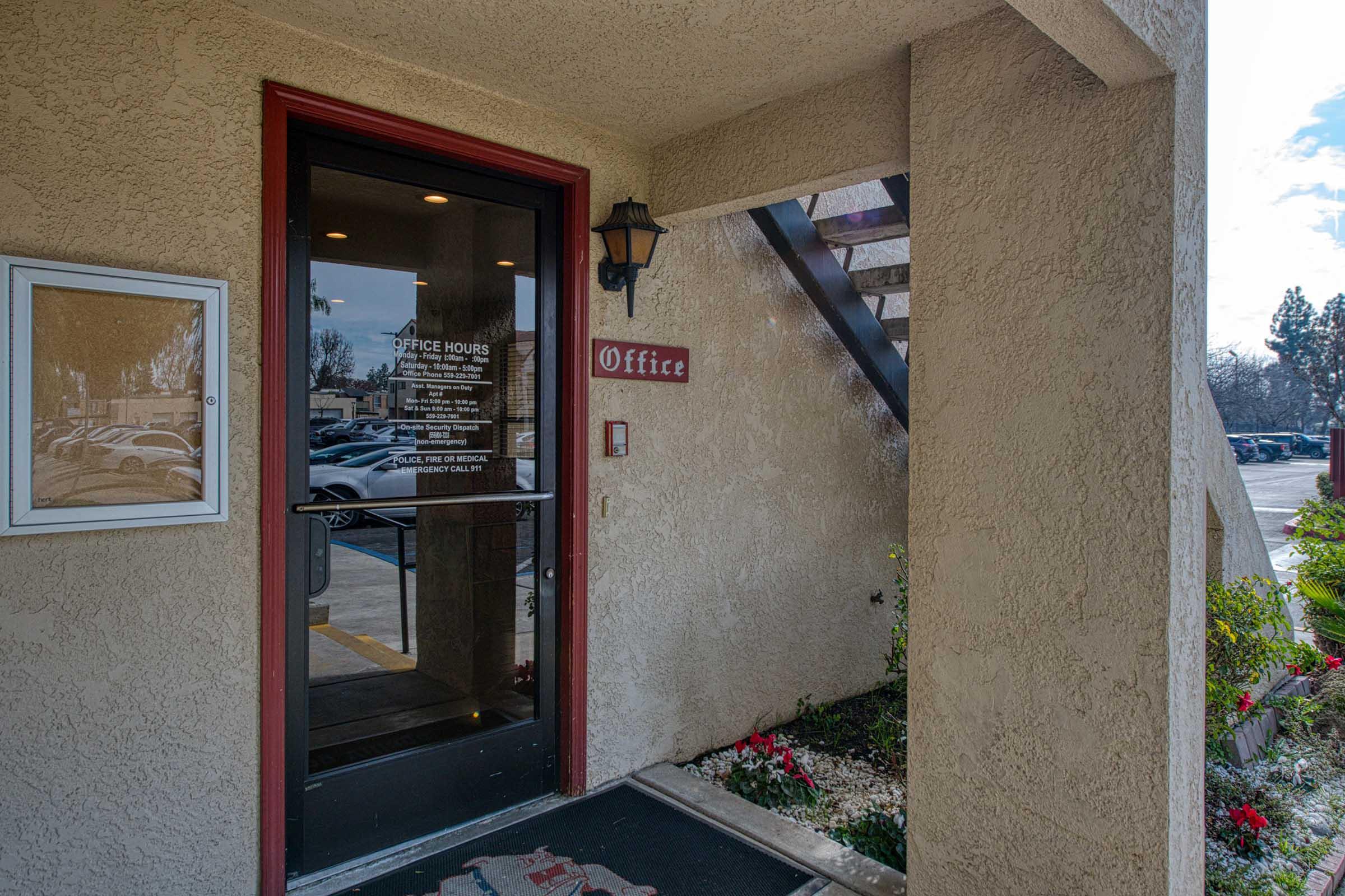 An entrance to an office building featuring a doorway with a sign that says "Office." There is a bulletin board to the left, a lantern-style light above the door, and a garden with flowers in front. The background shows parked cars and a cloudy sky.