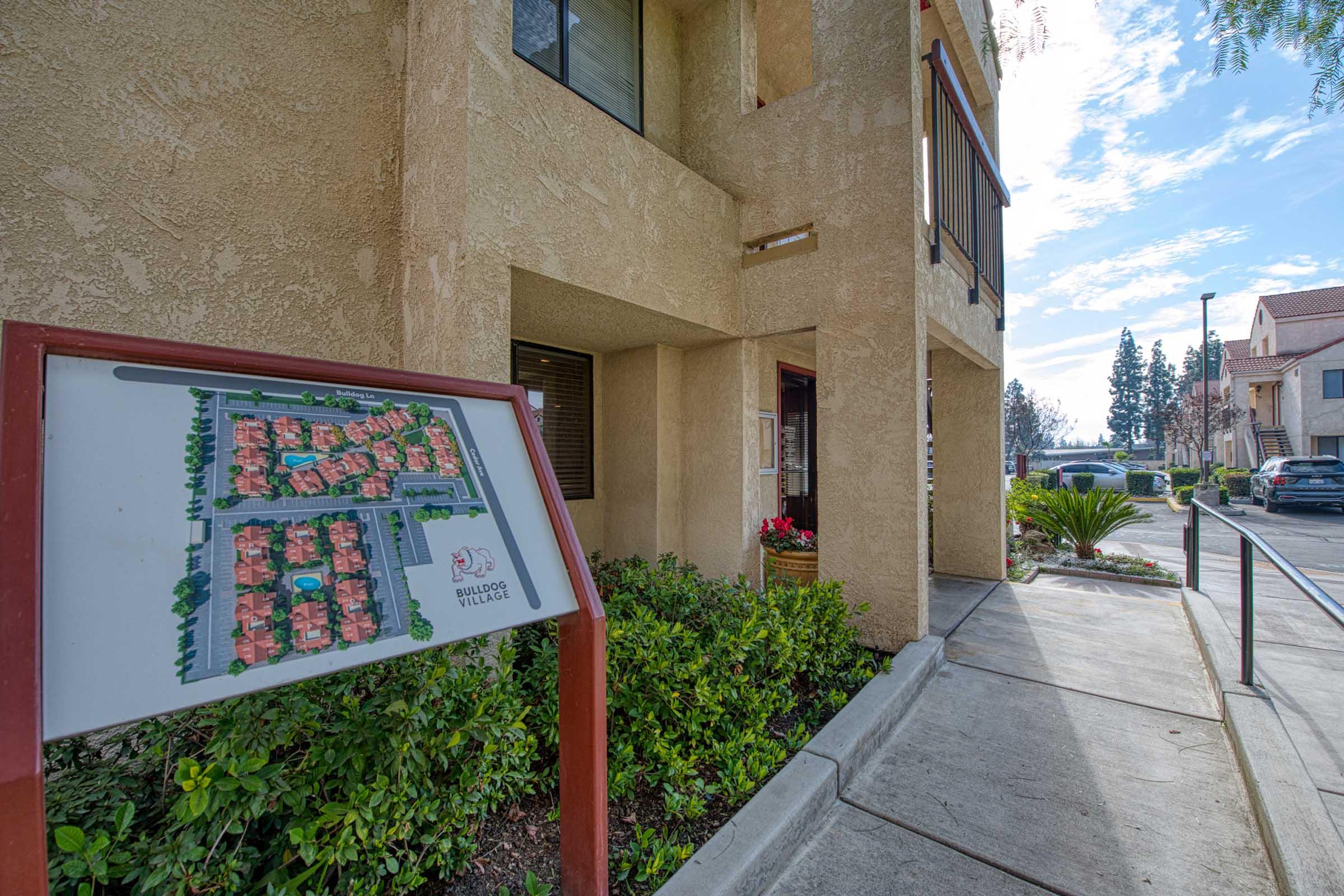 A view of an entrance to a building, featuring a sign with a layout map of the surrounding area. The pathway is lined with greenery, and there are decorative plants nearby. In the background, other buildings can be seen under a partly cloudy sky.