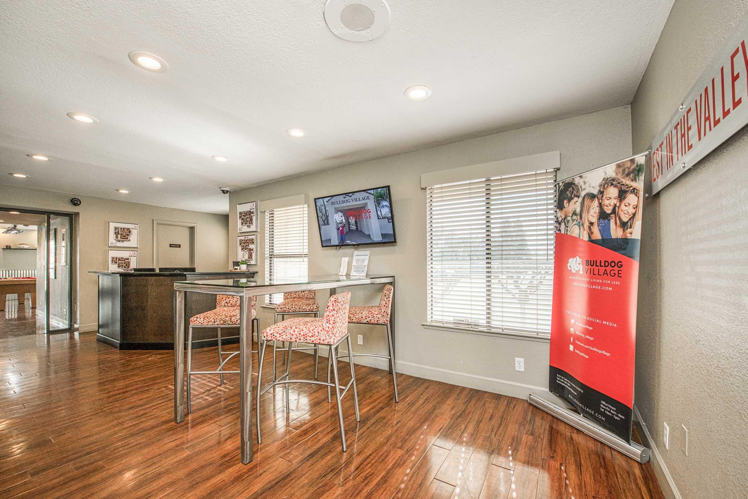 A modern common area featuring wooden floors, tall tables with patterned chairs, and a flat-screen TV mounted on the wall. A promotional banner for "Bulldog Village" is displayed, along with large windows allowing natural light into the space.