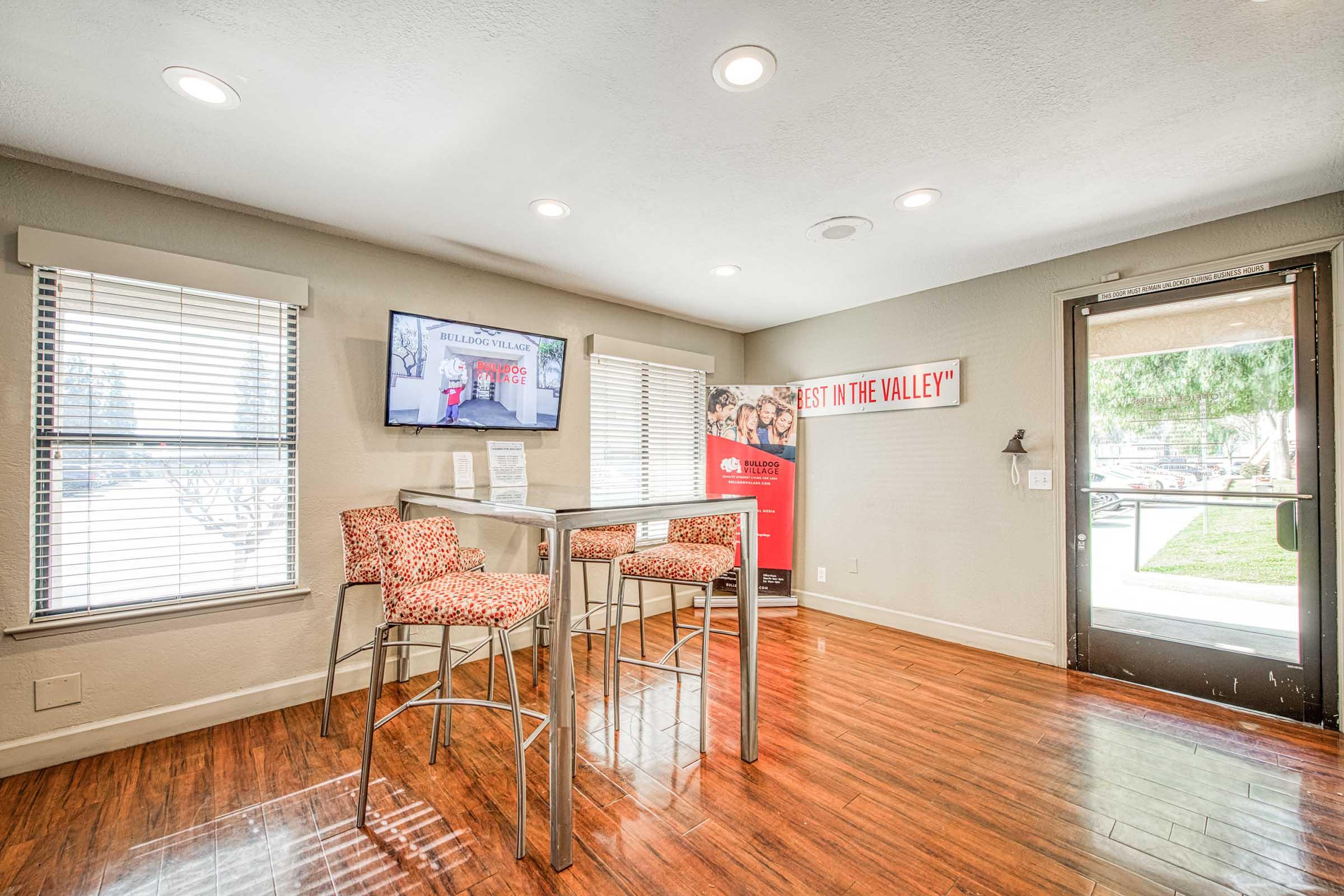 A bright and modern interior space featuring a tall table with four stylish chairs, large windows with blinds, and a TV mounted on the wall displaying a presentation. There is a banner in the corner that reads "BEST IN THE VALLEY." The flooring is polished wood, creating a welcoming environment.