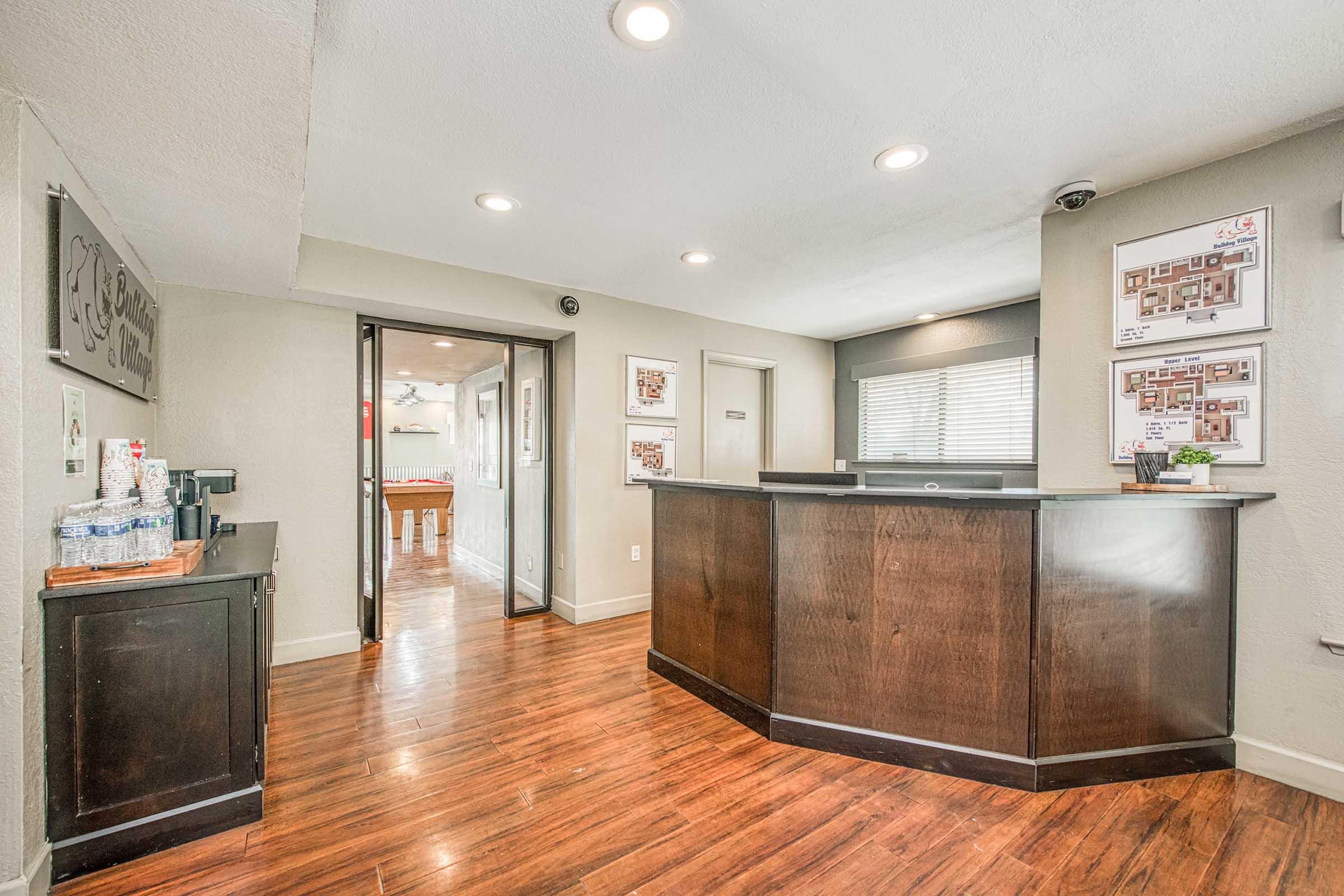 Interior view of a reception area featuring a wooden front desk, polished floors, and a small beverage station. The lighting is bright, with windows providing natural light. There are decorative photos on the walls and an open doorway leading to another room in the background.