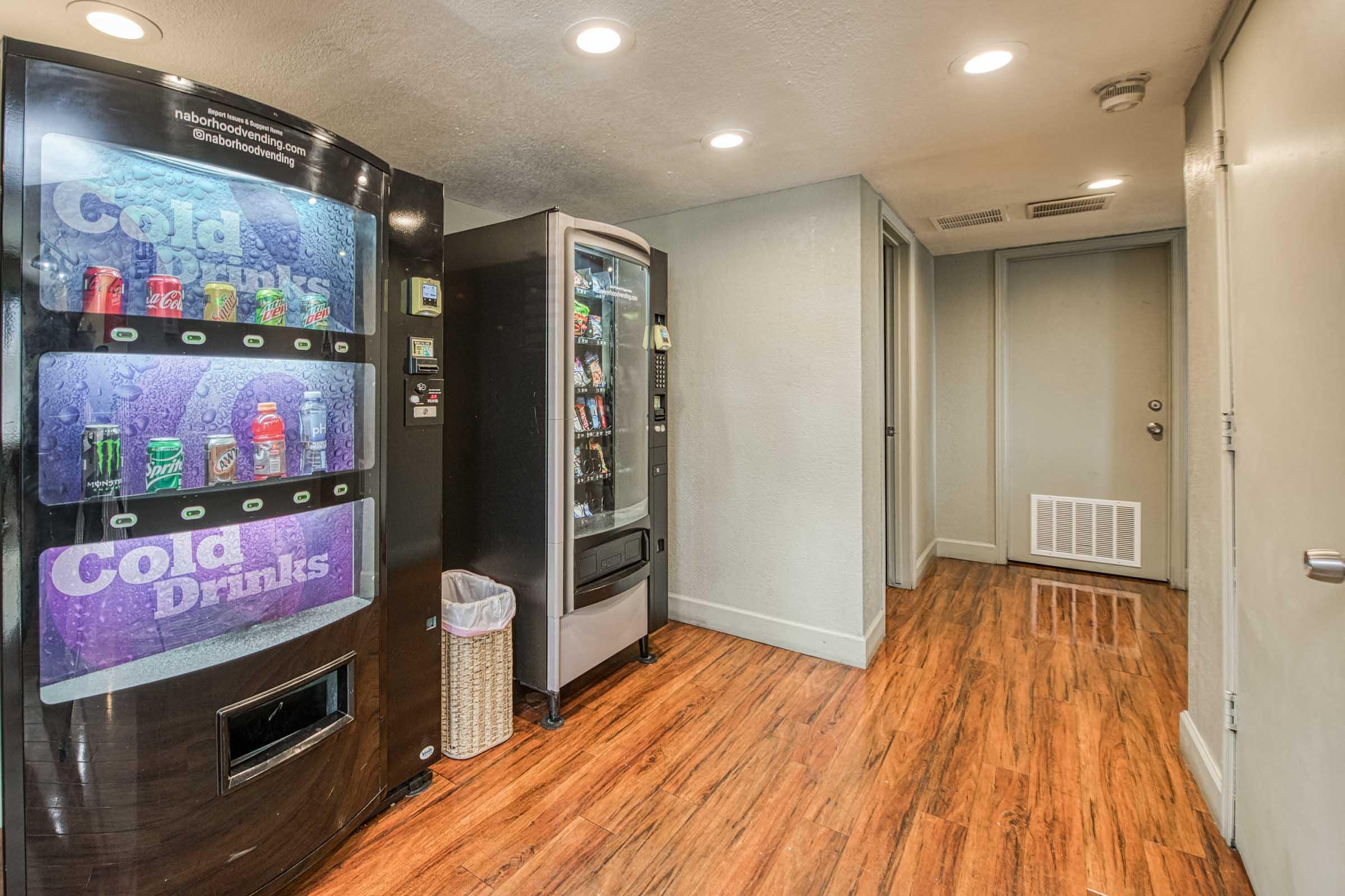 A hallway featuring two vending machines: one dispenses cold drinks, while the other offers snacks. The floor has a wooden finish, and there is a trash bin next to the drink machine. A closed door is visible at the far end of the hallway. Bright lighting enhances the clean, bright atmosphere.