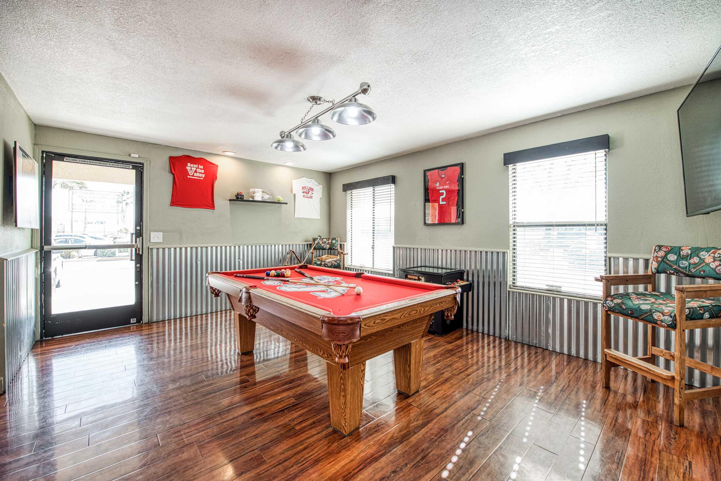 A billiards room featuring a red pool table under bright lights. The walls are decorated with framed jerseys and art. Large windows allow natural light, and there's a chair with a cushion nearby. The floor has a polished wood finish, creating a warm and inviting atmosphere.