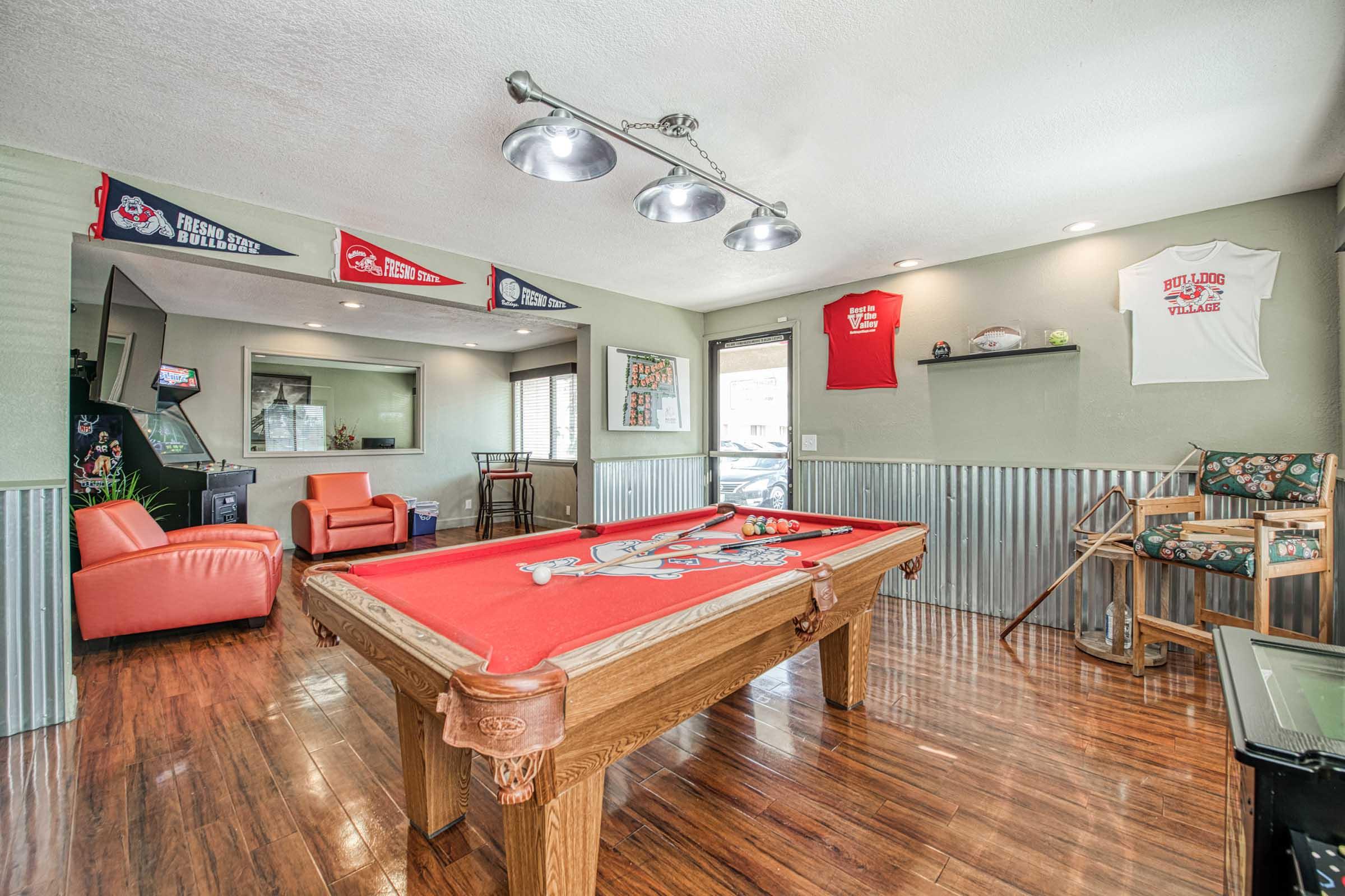 A cozy recreation room featuring a pool table with a red felt surface, wooden frame, and a rustic charm. The space includes orange leather chairs, sports-themed decorations, pennants on the walls, and a small arcade game console in the background. The flooring is polished wood, adding warmth to the room.