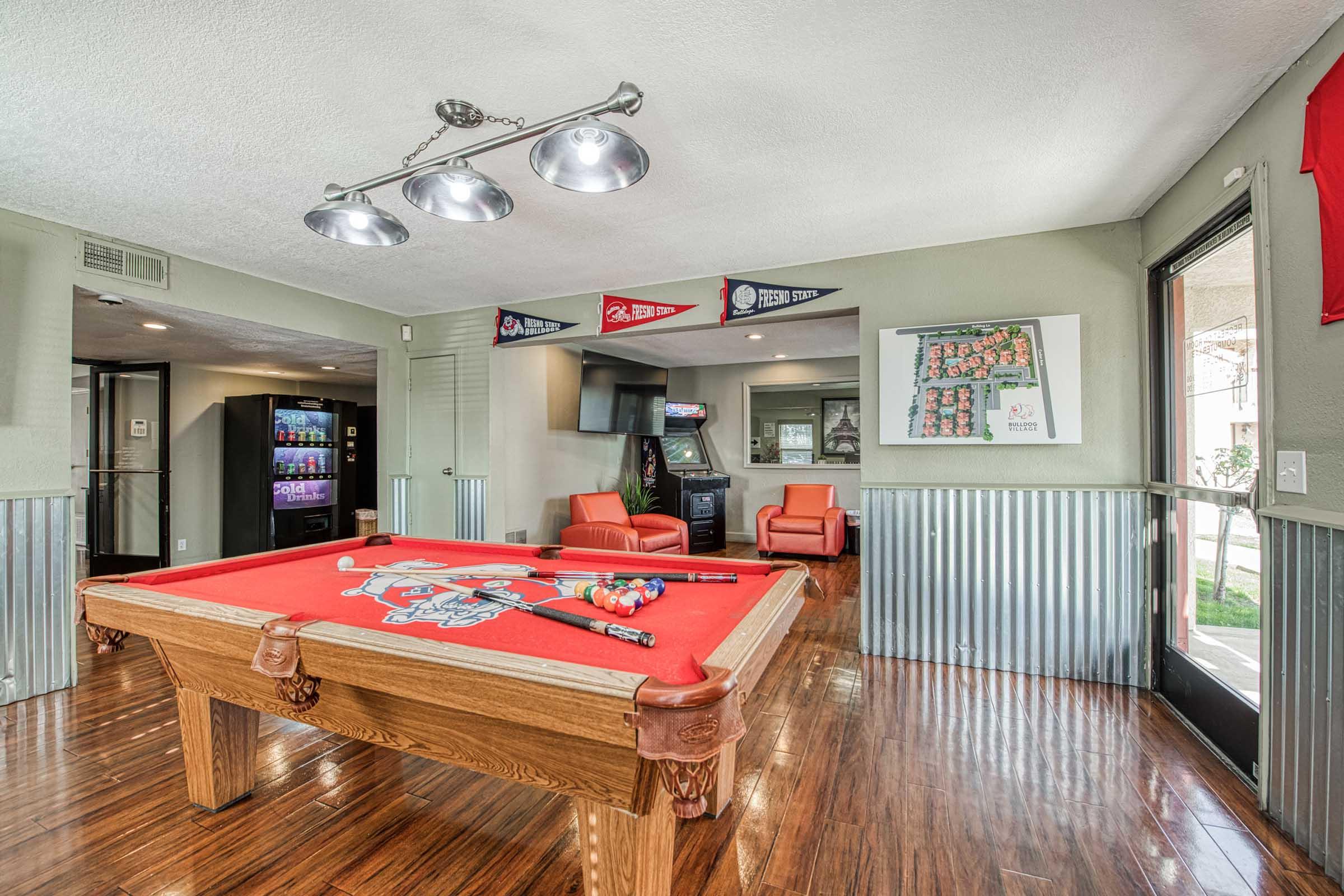 A spacious game room featuring a red felt pool table with cues and balls ready to play. Modern lighting hangs above the table. In the background, there are vending machines, a television, and arcade games. The walls are decorated with sports pennants. Large windows provide natural light, enhancing the inviting atmosphere.