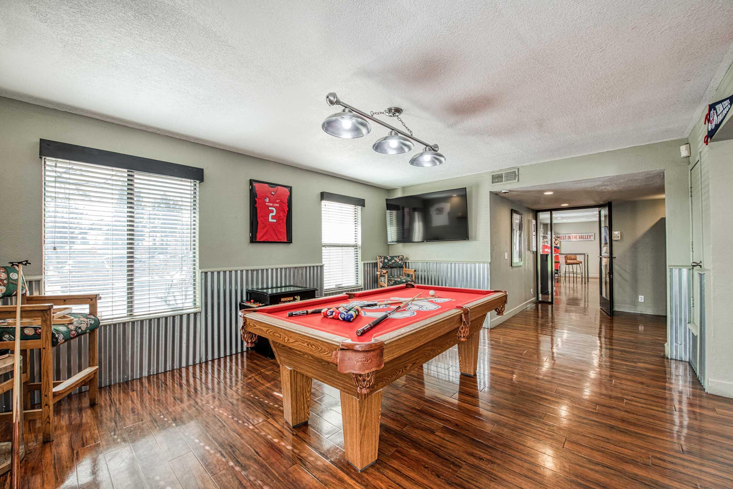 A bright, spacious game room featuring a red pool table in the center, surrounded by hardwood flooring. Large windows provide natural light, and there are wall-mounted lights above the table. The room includes a flat-screen TV and decor items, creating a cozy and inviting atmosphere for entertainment.