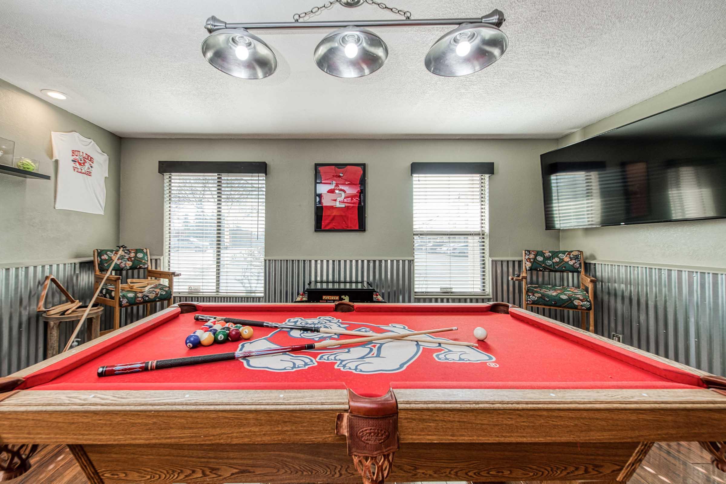 A well-lit game room featuring a red pool table with a Bulldog logo, cue sticks, and colorful billiard balls. The walls are decorated with sports memorabilia and two windows allow natural light. There are two comfortable chairs and a flat-screen TV on one wall.
