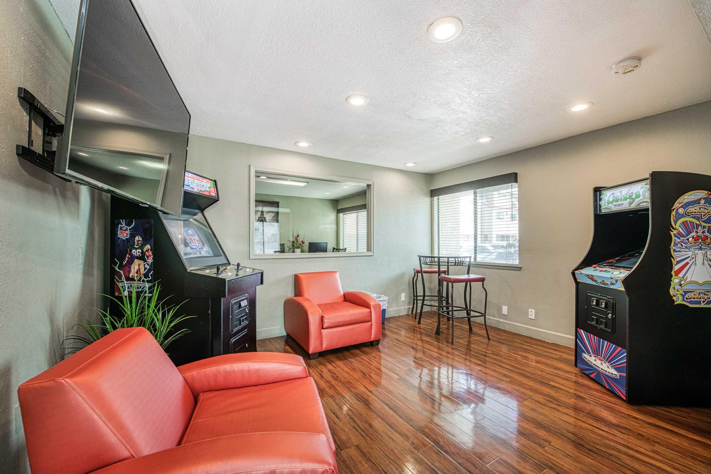 A cozy game room featuring two arcade machines, a large flat-screen TV mounted on the wall, and a high-top bar table with stools. The space has natural light coming through the windows and is furnished with two modern orange armchairs, complemented by wooden flooring.