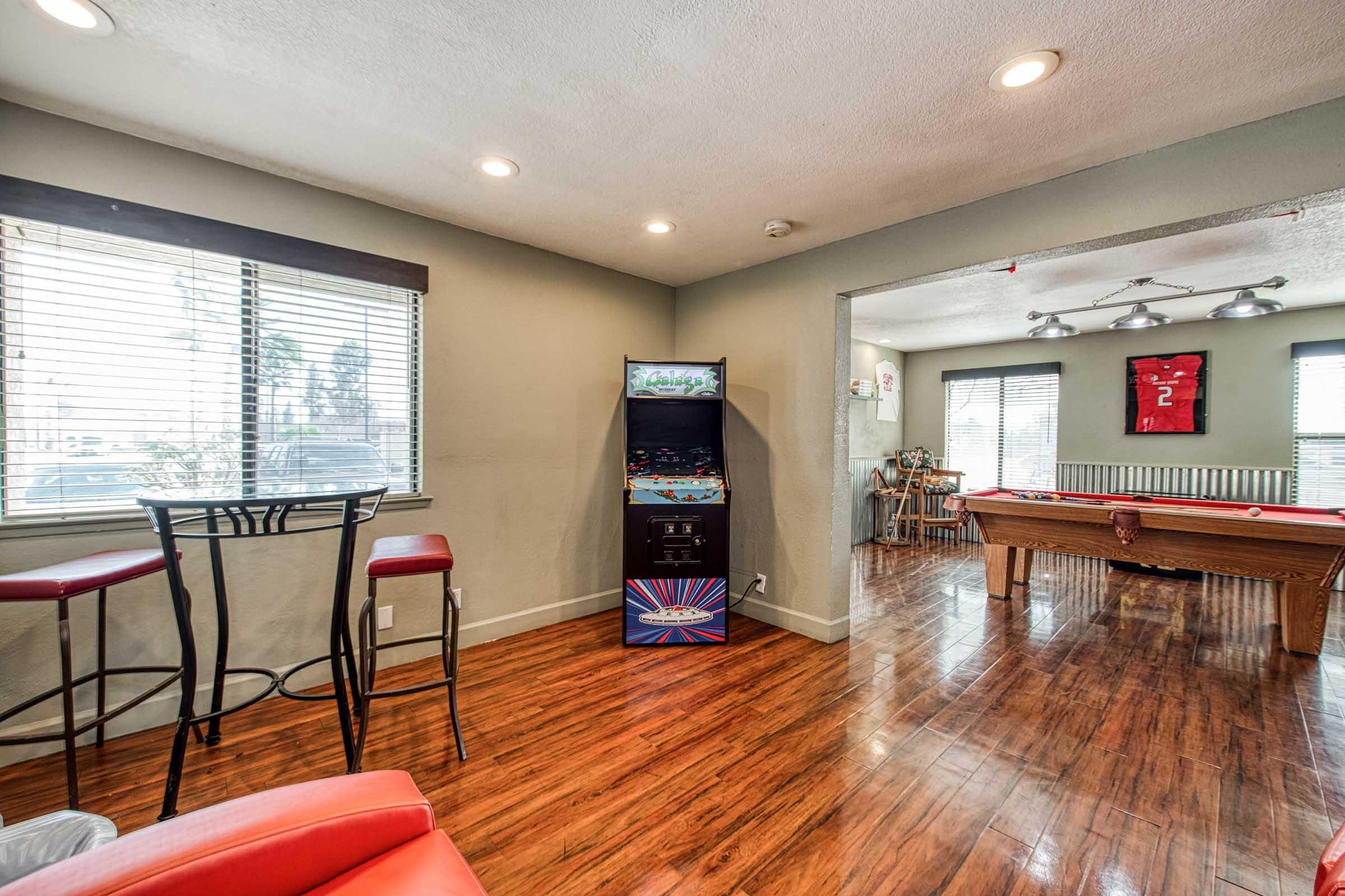 A modern living space featuring a cozy seating area with red chairs, a small bar table, an arcade game machine, and a pool table. Large windows allow natural light to fill the room, highlighting the hardwood floor and contemporary decor. The atmosphere is inviting and stylish.