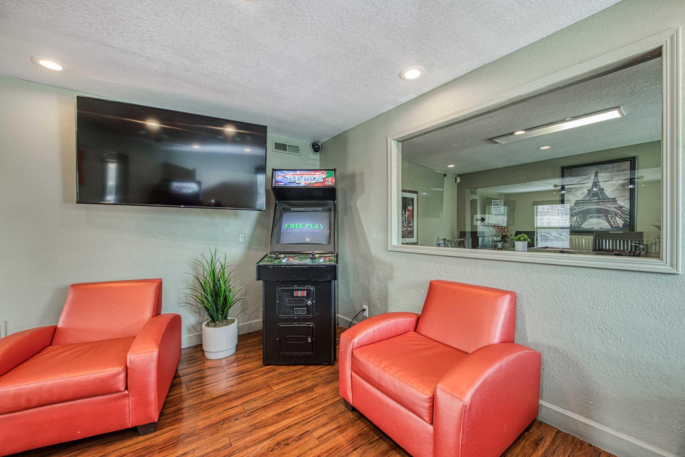 A cozy lounge area featuring two red chairs, a television mounted on the wall, and an arcade game cabinet that offers free play. A potted plant adds a touch of greenery to the modern decor. There’s a large mirror reflecting the space, enhancing the room’s openness and inviting atmosphere.
