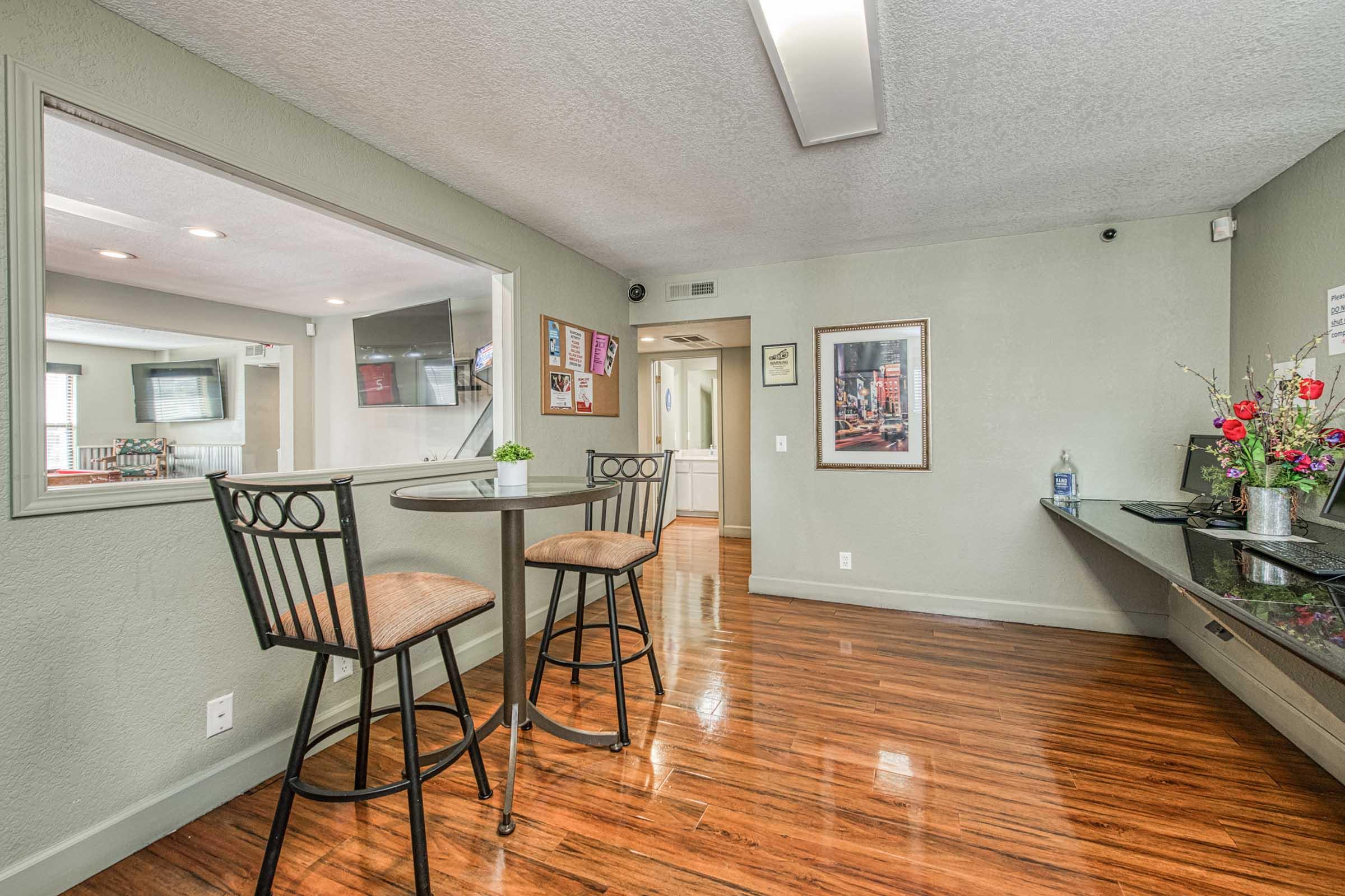 A small room featuring a round table with two bar stools, wooden flooring, and green walls. There's a framed picture on the wall, a decorative plant on the table, and a side table with flowers and a water dispenser. A large window allows natural light and offers a view of another area.