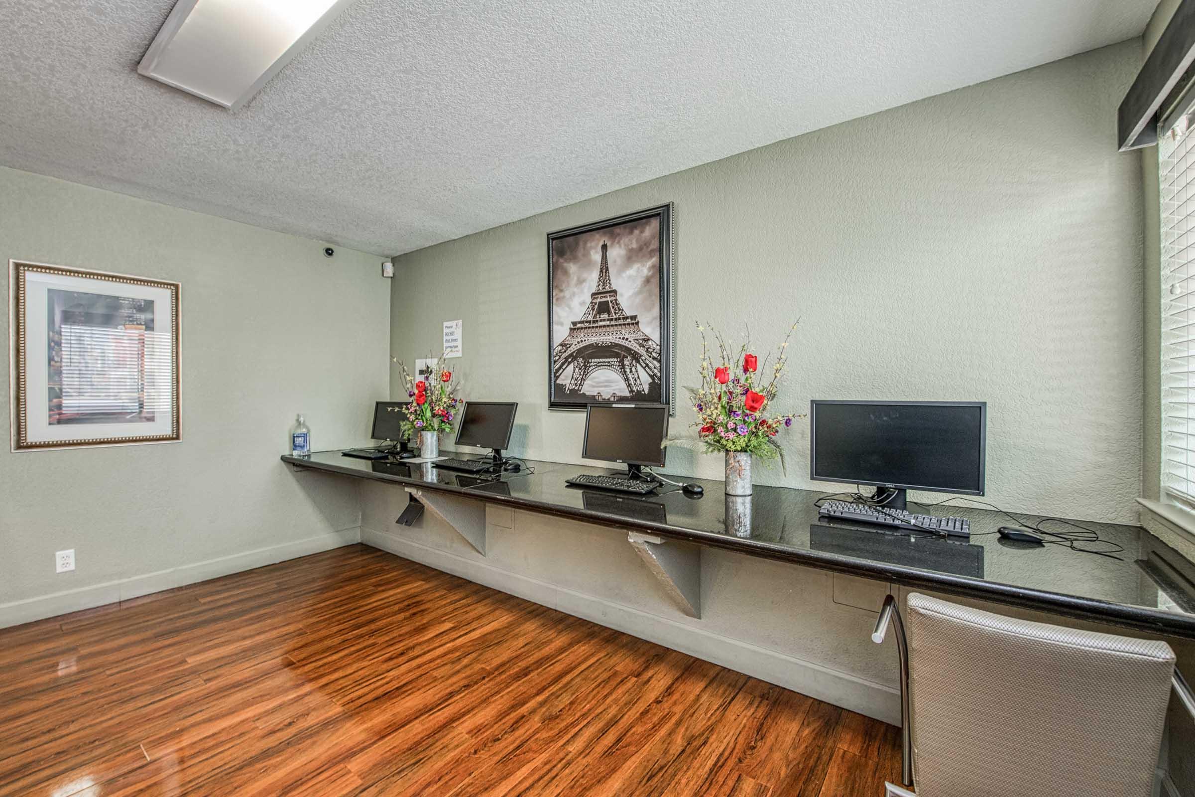 A modern workspace featuring a long desk with three computers and stylish chairs. The walls are painted a soft green, adorned with an Eiffel Tower photograph and decorative vases with flowers. The flooring is a polished wooden surface, creating a welcoming and professional atmosphere.