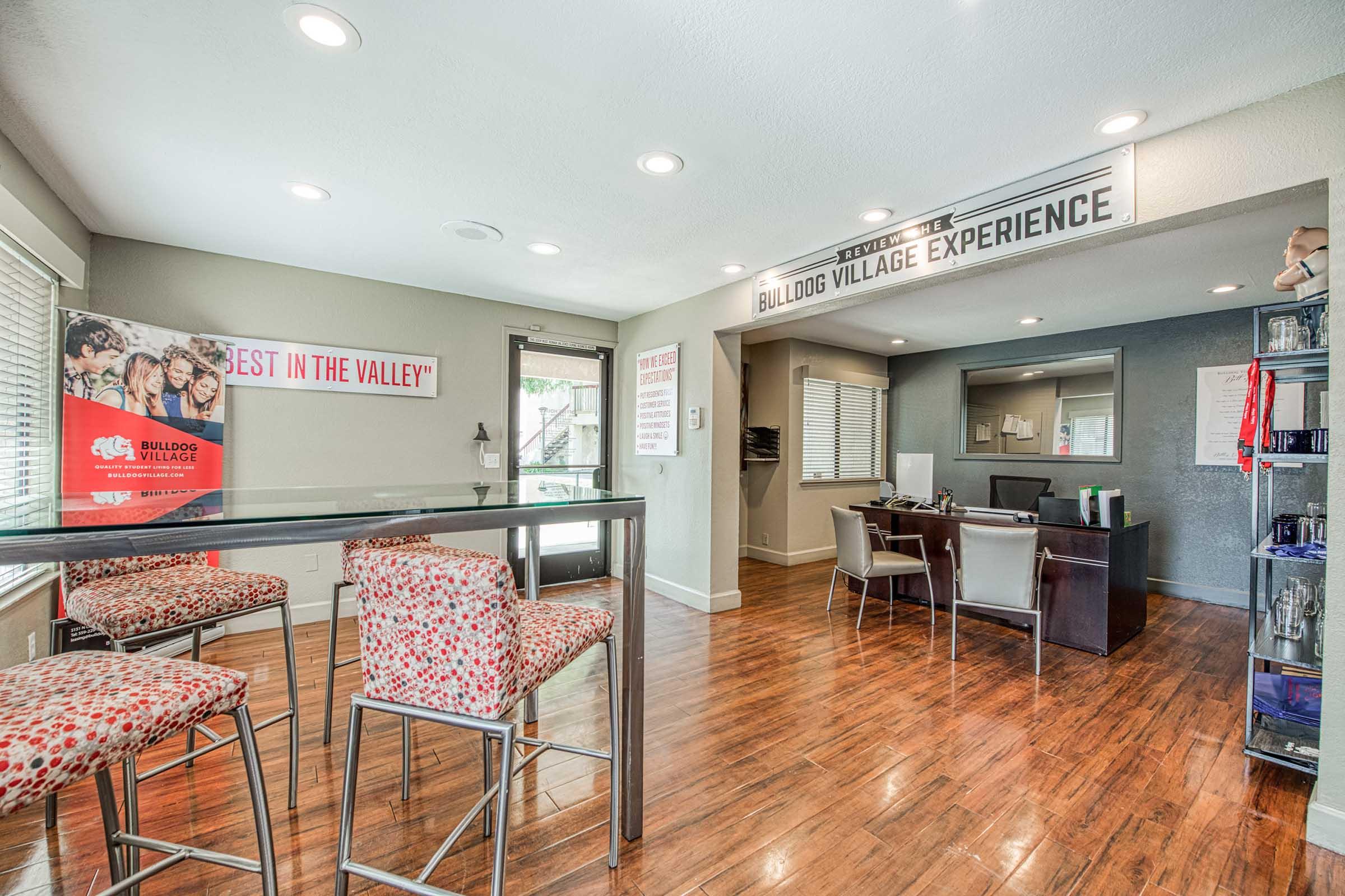 Interior view of a welcoming reception area, featuring a modern design with wooden floors, a desk for inquiries, and seating at high bar tables. Promotional signage for "Bulldog Village" is visible, creating a friendly atmosphere for residents or visitors. Natural light enhances the inviting space.