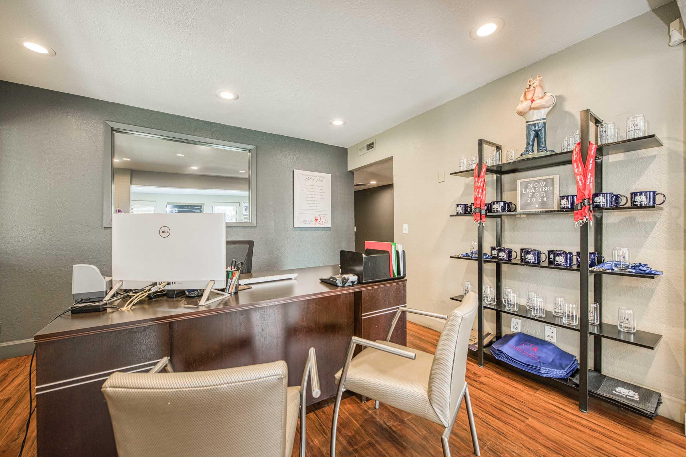 A modern office reception area featuring a dark wood desk with a computer, two chairs, and wall-mounted shelves displaying mugs and glassware. The walls are light gray, with decorative items and a mirror. Bright overhead lighting creates a welcoming atmosphere.