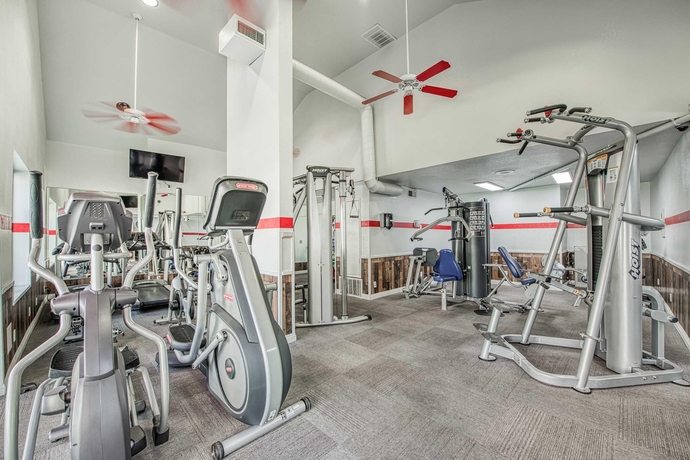 A modern gym interior featuring various exercise equipment, including elliptical machines, weight machines, and a stationary bike. The space has a high ceiling with ceiling fans, red accents on the walls, and carpeted flooring, creating a bright and inviting workout environment.
