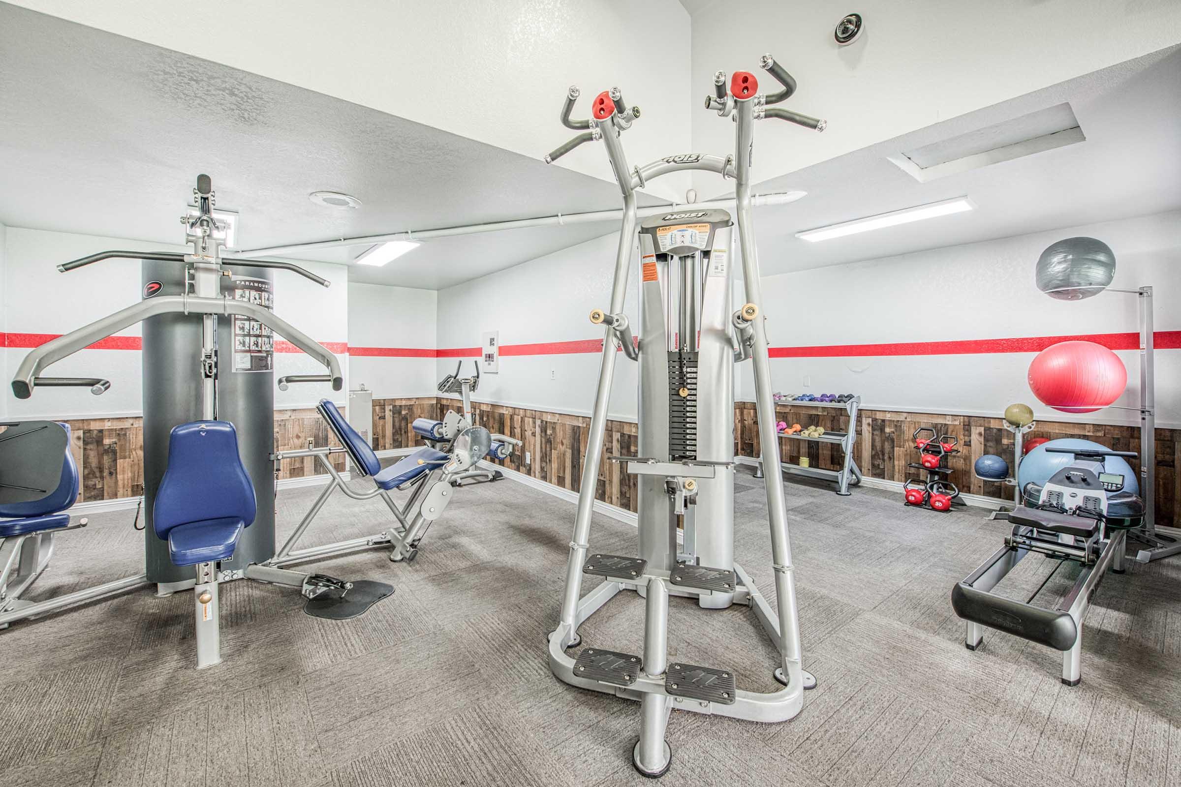A well-equipped gym interior featuring various fitness machines, including a multi-station weight machine, a leg press, and free weights. The walls are painted in light colors with a red accent, and there are exercise balls and resistance bands neatly organized, creating an inviting workout space.