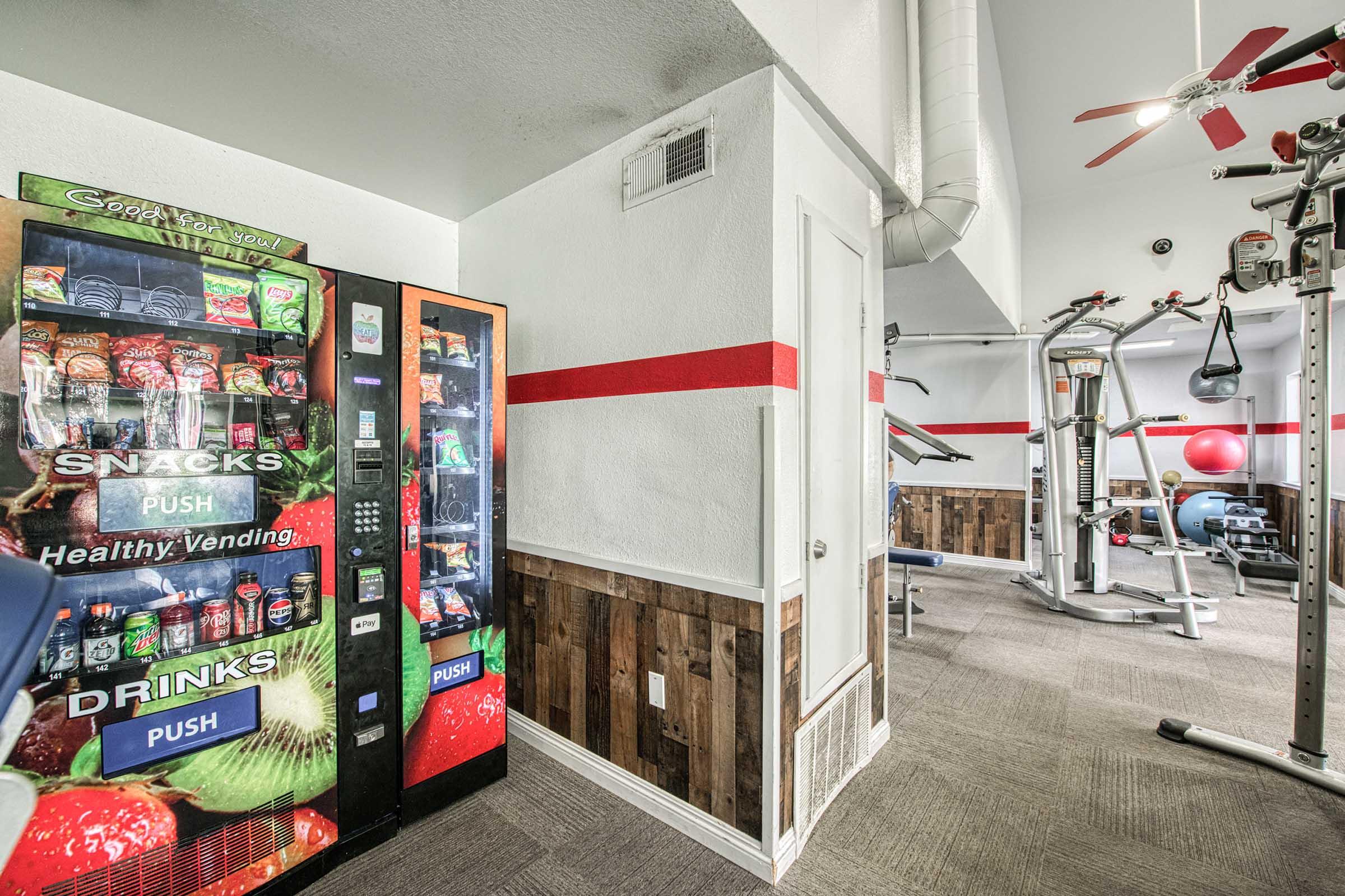 Vending machine displaying snacks and drinks in a fitness center, with a gym area visible in the background featuring exercise equipment and workout space. The walls are decorated with a red stripe and wood paneling.