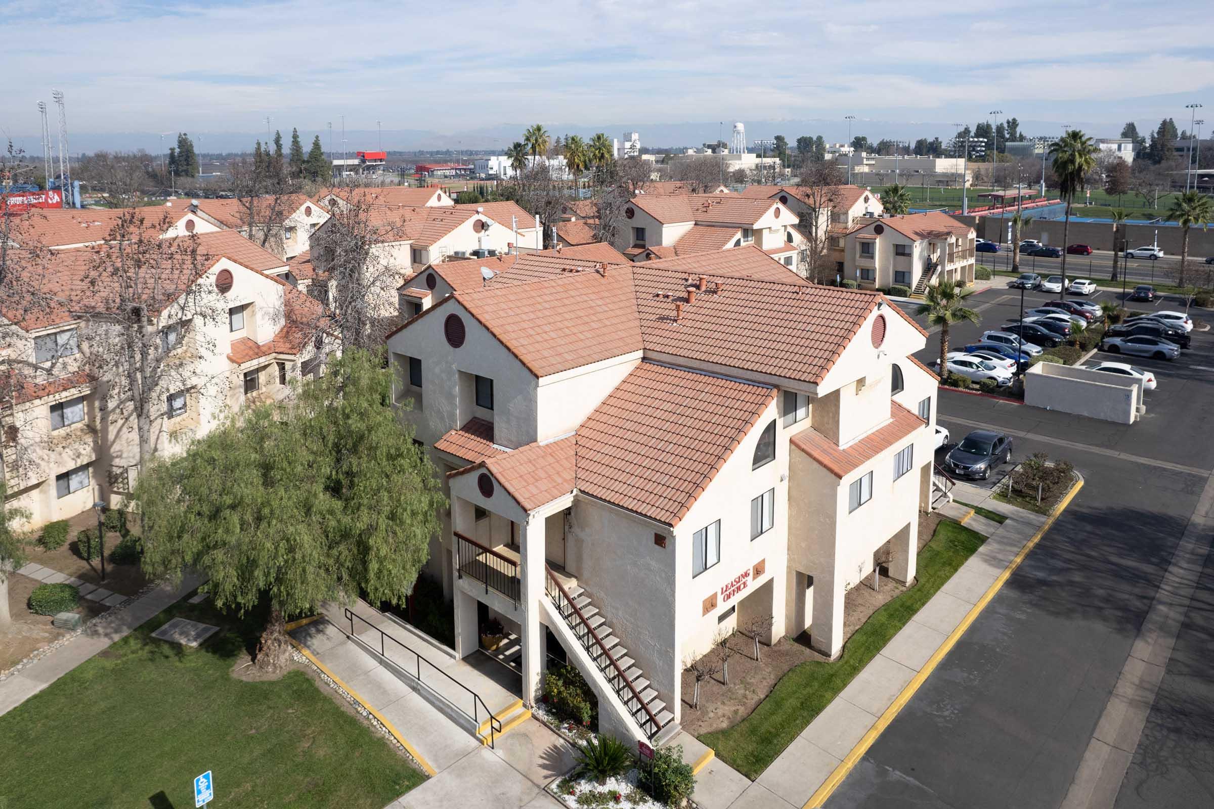 Aerial view of a residential complex with light-colored buildings featuring red-tiled roofs. A green lawn with a tree is in the foreground, and parked cars line the side of the building. In the background, there are sports fields and distant hills.