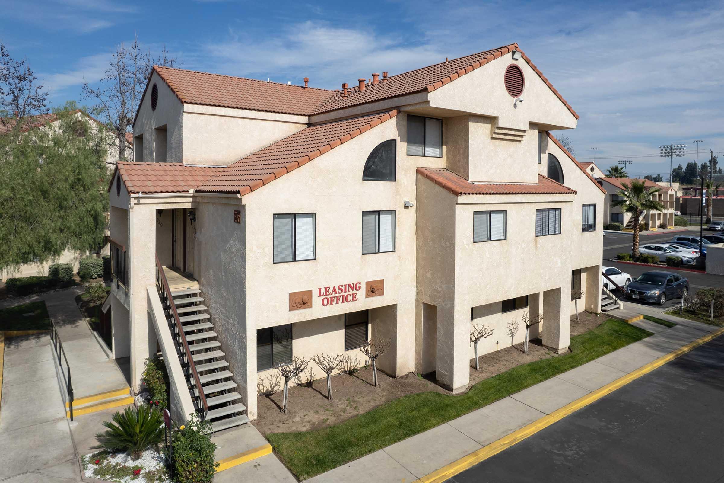 A two-story building with a beige exterior featuring a leasing office sign. There are several windows on each floor, and a staircase leading to the second level. Surrounding the building are neatly trimmed shrubs and a paved parking lot visible in the foreground. The sky is clear with a few clouds.