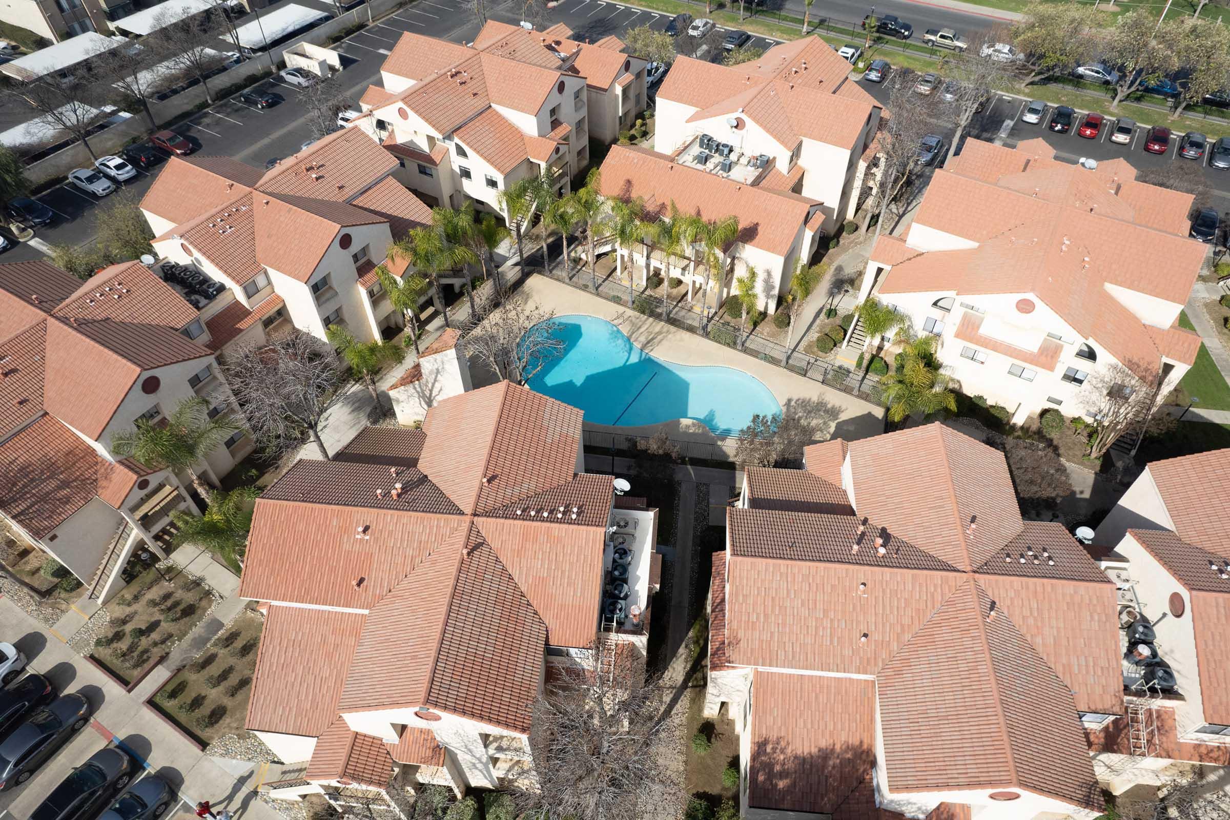Aerial view of a residential complex featuring several buildings with red tiled roofs surrounding a central swimming pool. Palm trees are visible near the pool area, and parking spaces are lined up in the background. The scene captures a sunny day, showcasing a well-maintained community.
