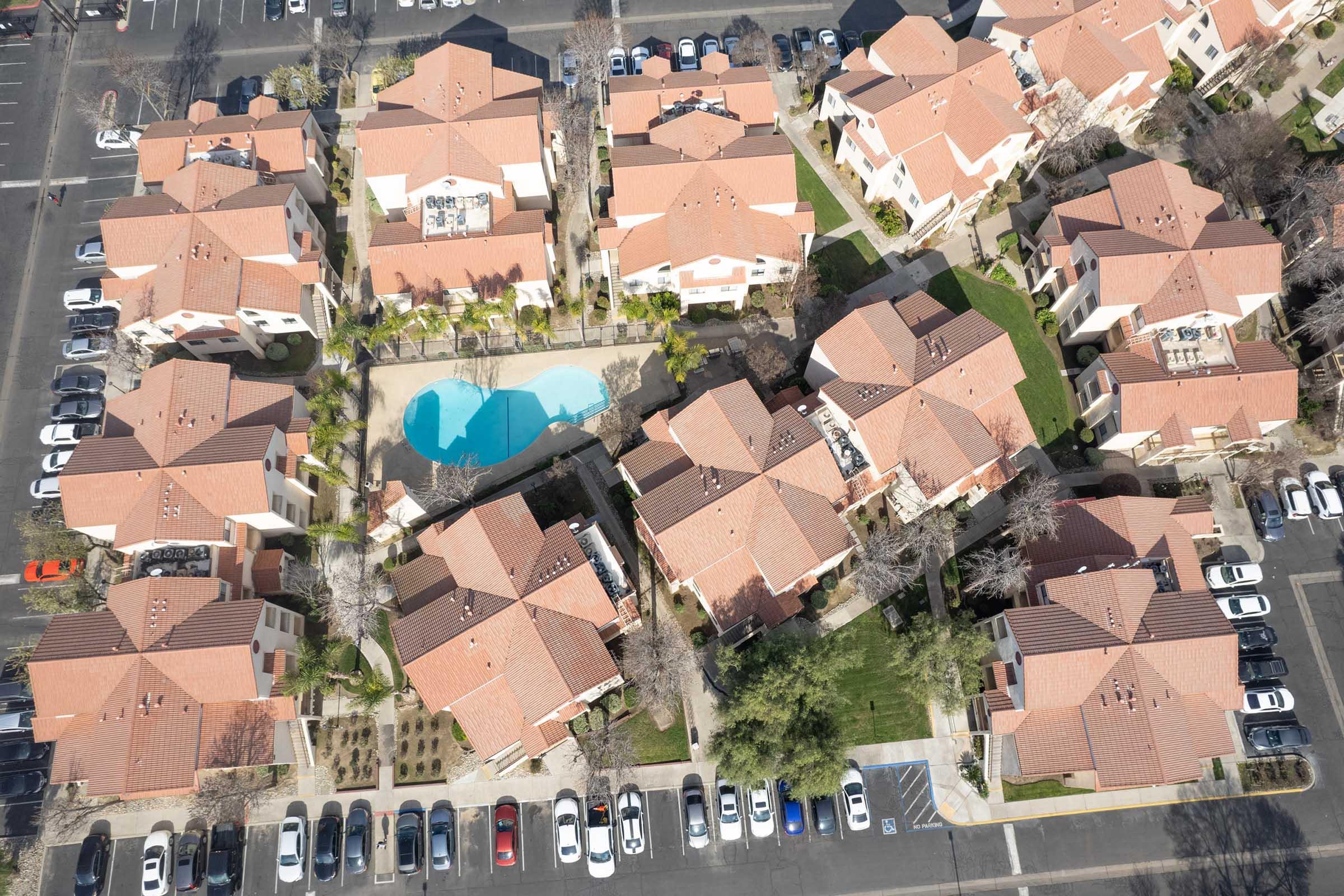 Aerial view of a residential complex featuring multiple buildings with orange rooftops. The layout includes a central pool surrounded by landscaped areas. Surrounding parking spaces are filled with various cars, and nearby greenery adds to the community atmosphere.