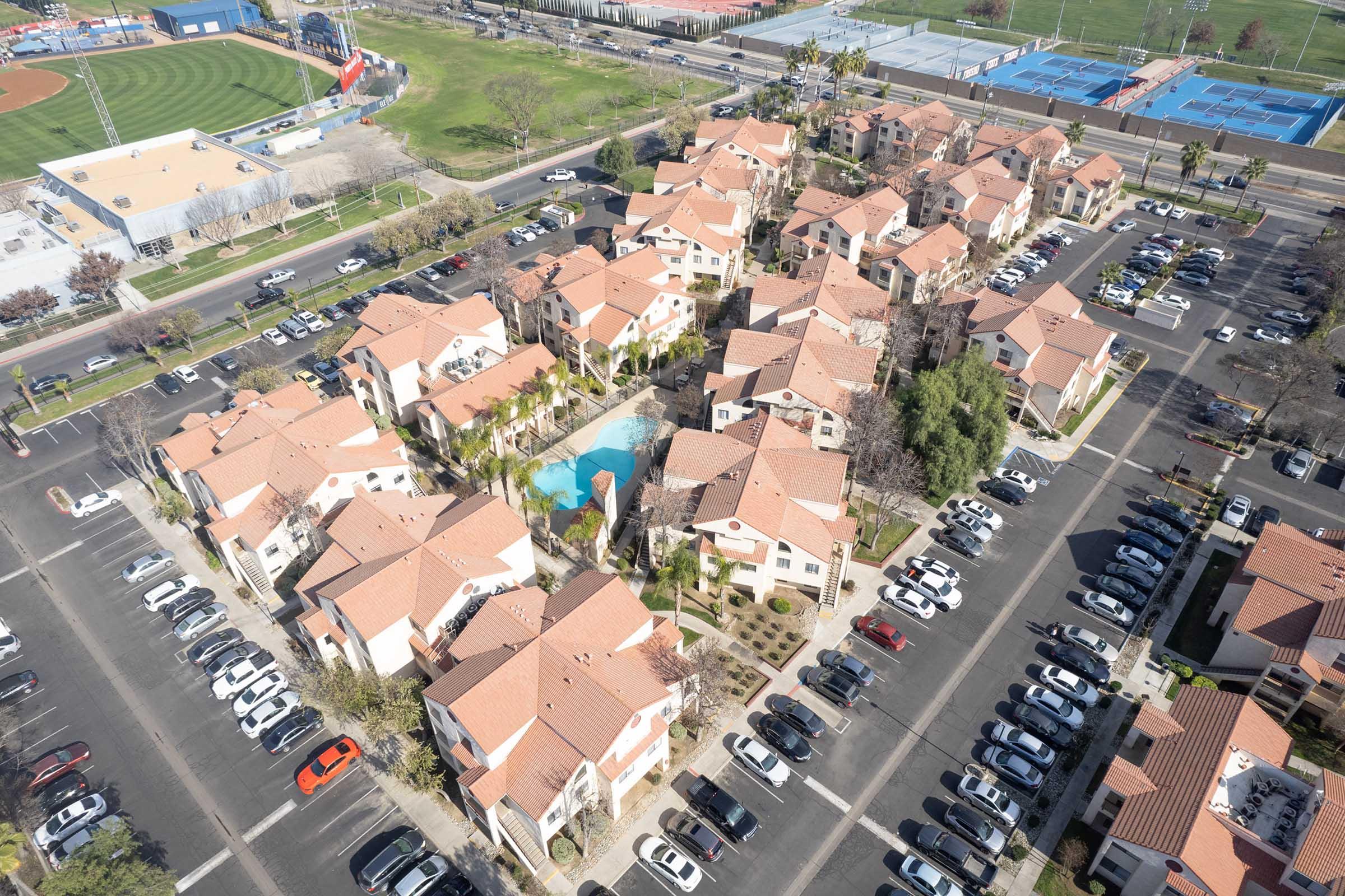 Aerial view of a residential complex featuring several residential buildings with terracotta roofs, a central pool area surrounded by palm trees, and a large parking lot filled with parked cars. Nearby sports fields and a baseball stadium are visible in the background.