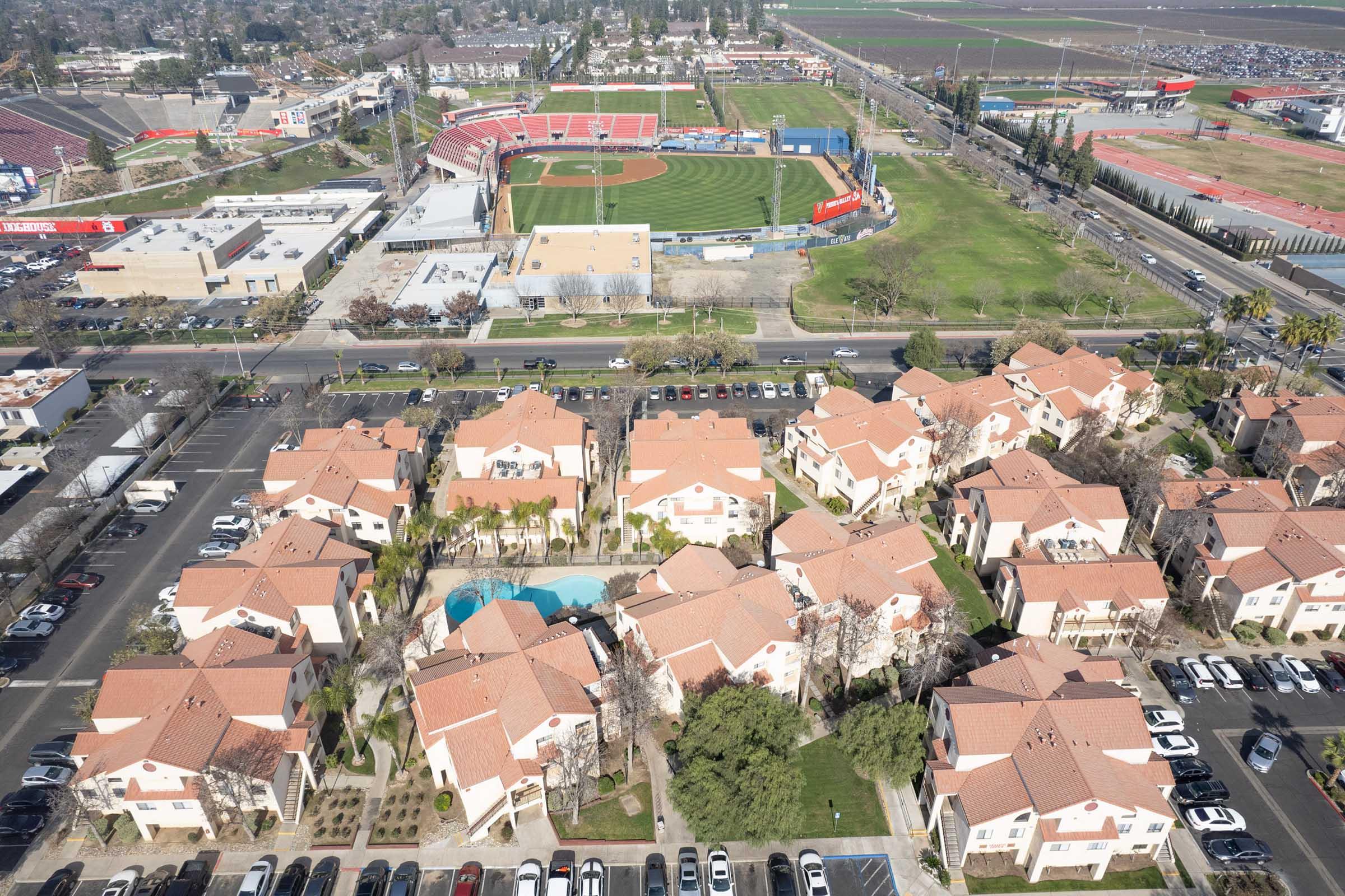 Aerial view of a residential area featuring several buildings with red-tiled roofs, a central pool, and parking spaces. In the background, a baseball field and sports facilities are visible, surrounded by green fields and a cityscape. The image captures a blend of urban living and recreational space.