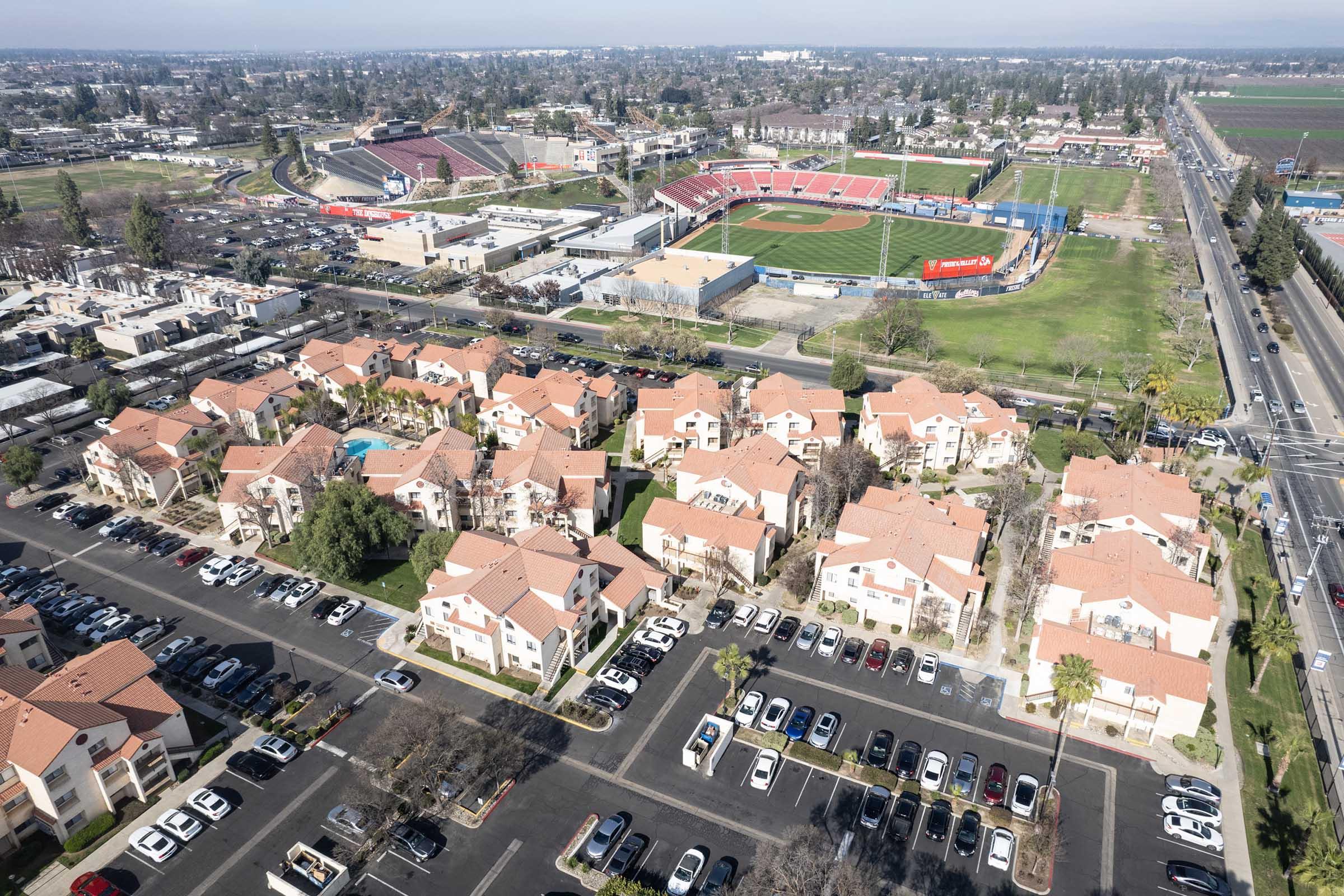 Aerial view of a residential area featuring multiple apartment buildings with orange roofs, surrounded by parking lots. In the background, a baseball field and sports complex are visible, along with green fields and a clear sky. The scene captures a blend of urban living and recreational spaces.