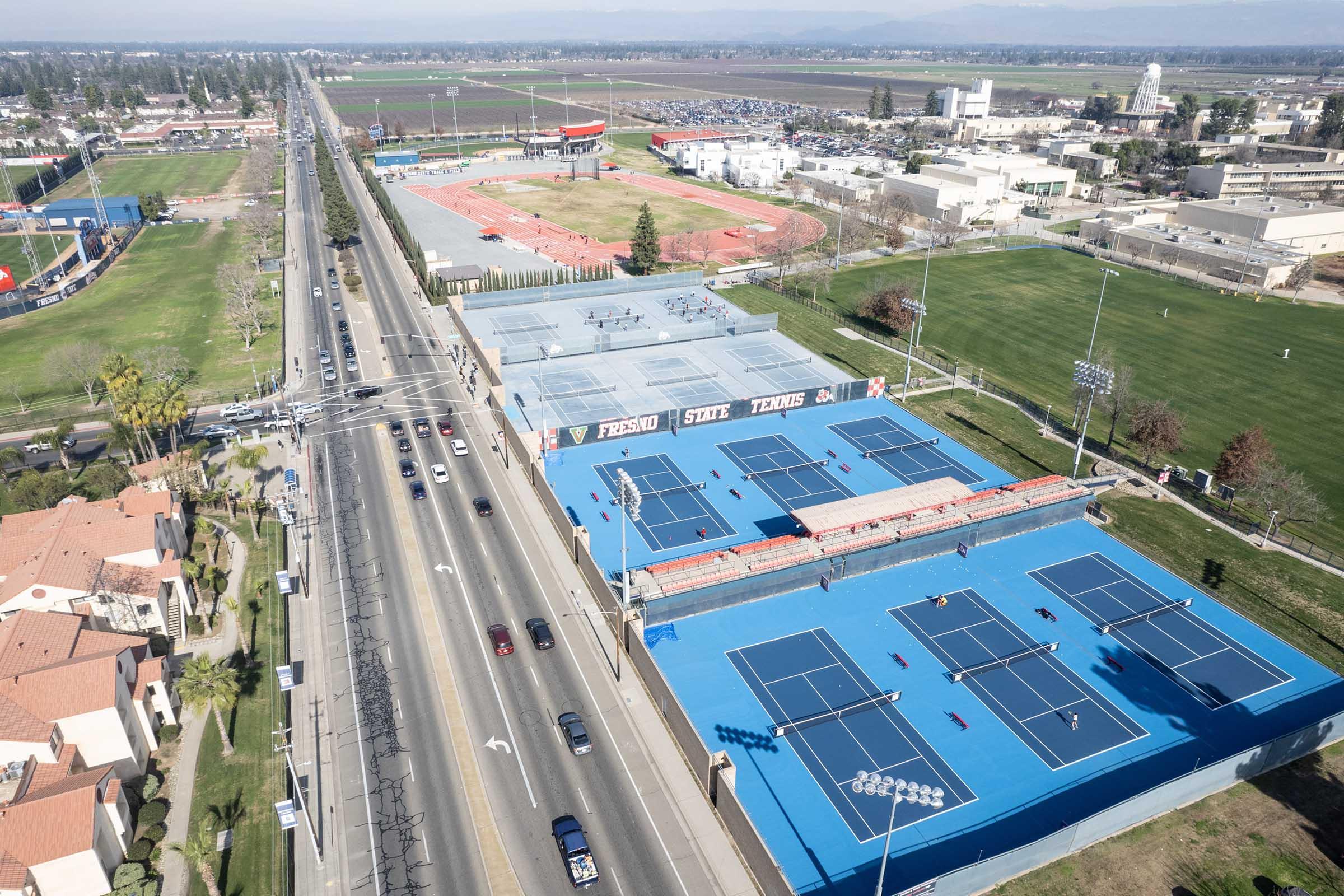 Aerial view of a sports complex featuring bright blue tennis courts, adjacent to a road. Nearby, there are several fields and a sports facility, with a small stadium visible. The area is surrounded by trees and open fields, showcasing a sunny day.