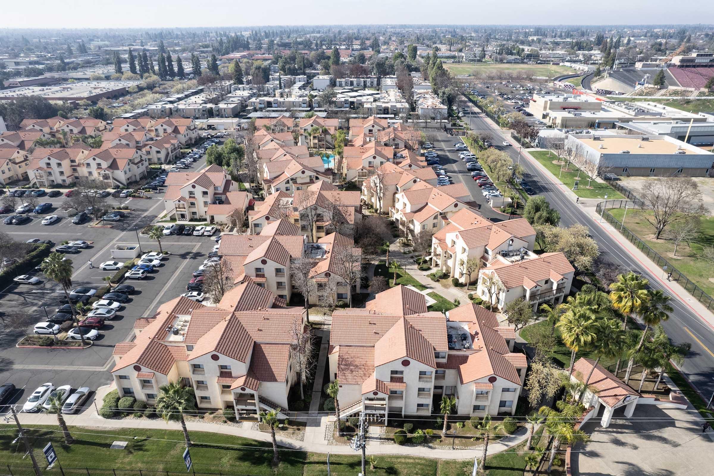 Aerial view of a residential complex featuring multiple buildings with red roofs surrounded by green landscaping and palm trees. The area includes parking lots and is situated near urban development, with additional buildings and roads visible in the background.
