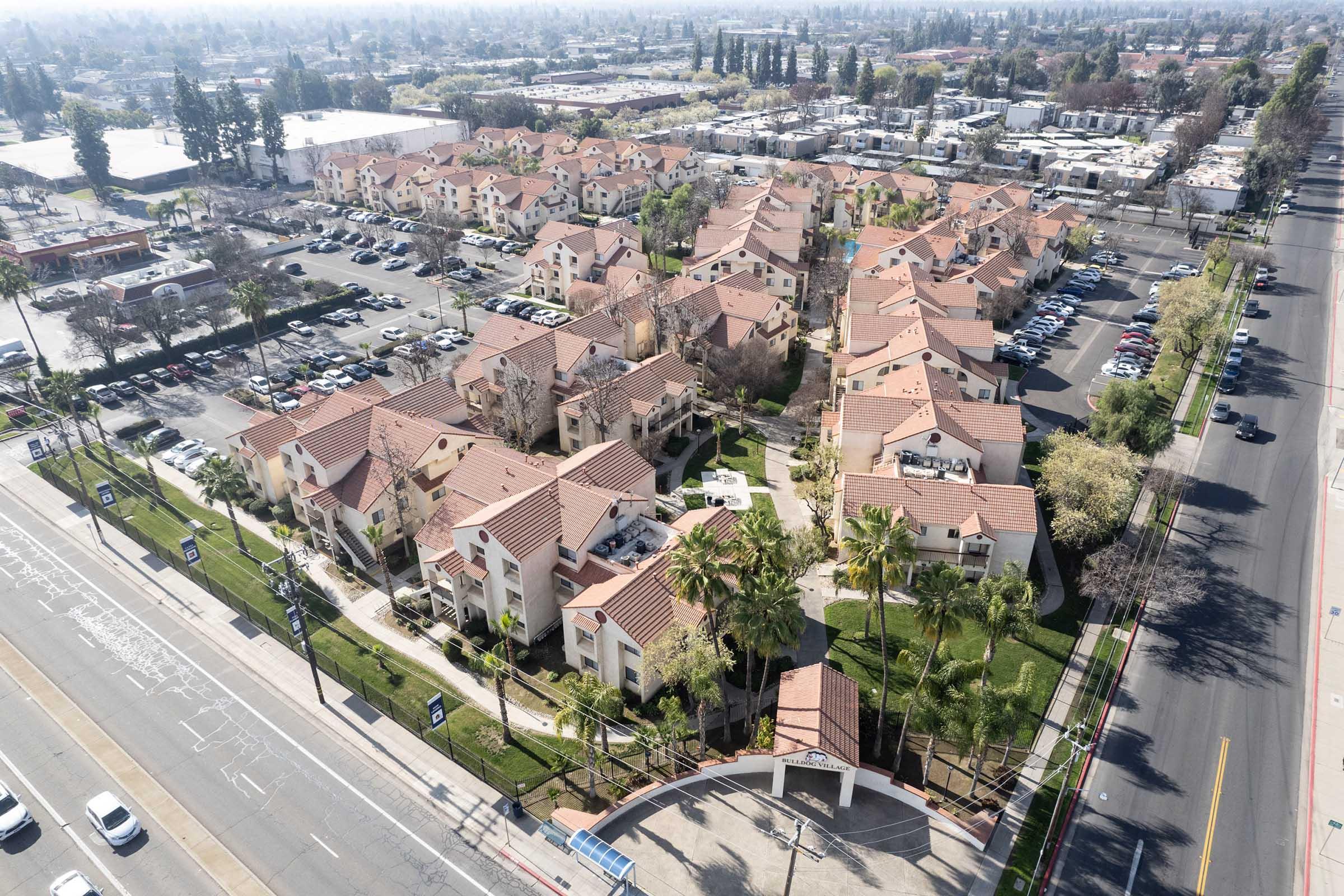 Aerial view of a residential area with several multi-story buildings featuring red-tiled roofs. Surrounding the buildings are green spaces and palm trees. In the background, there are additional structures and a parking lot filled with cars. A road runs alongside the property.