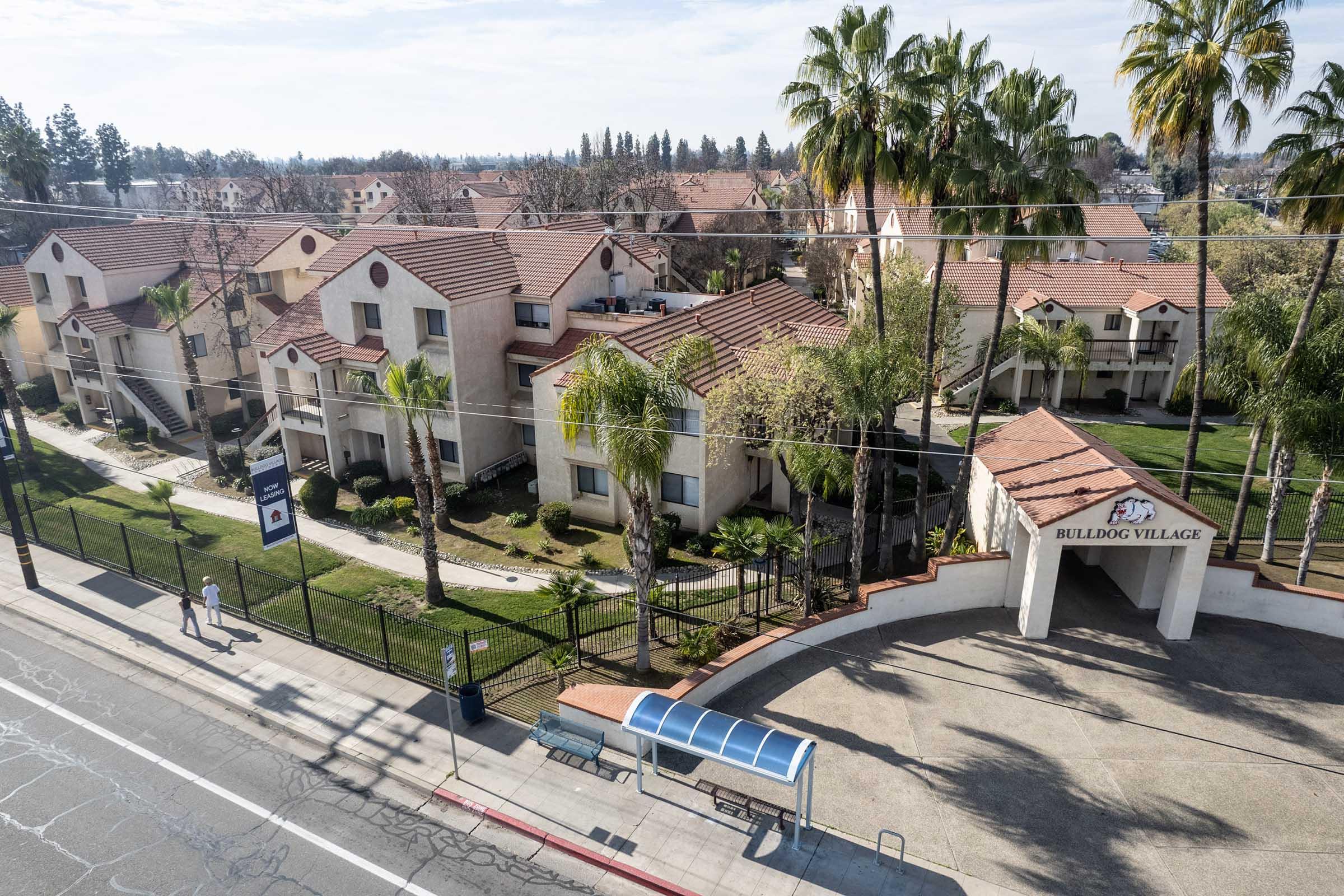 Aerial view of a residential complex named BullDog Village, featuring two-story buildings with red-tiled roofs and palm trees. A sidewalk runs alongside the complex, with a bus stop shelter in the foreground and a clear blue sky above. The area appears well-maintained and landscaped.