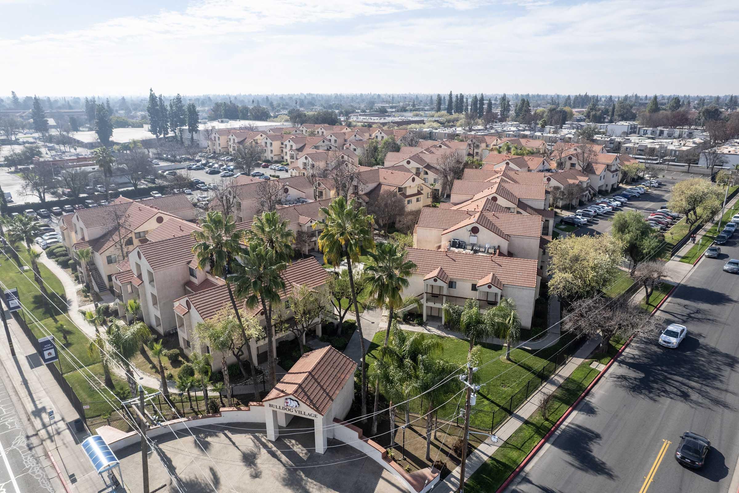 Aerial view of a residential area with multiple buildings featuring red tile roofs. The scene includes palm trees and well-maintained green spaces, with a road visible in the foreground. In the background, additional homes and parking lots can be seen against a clear sky.
