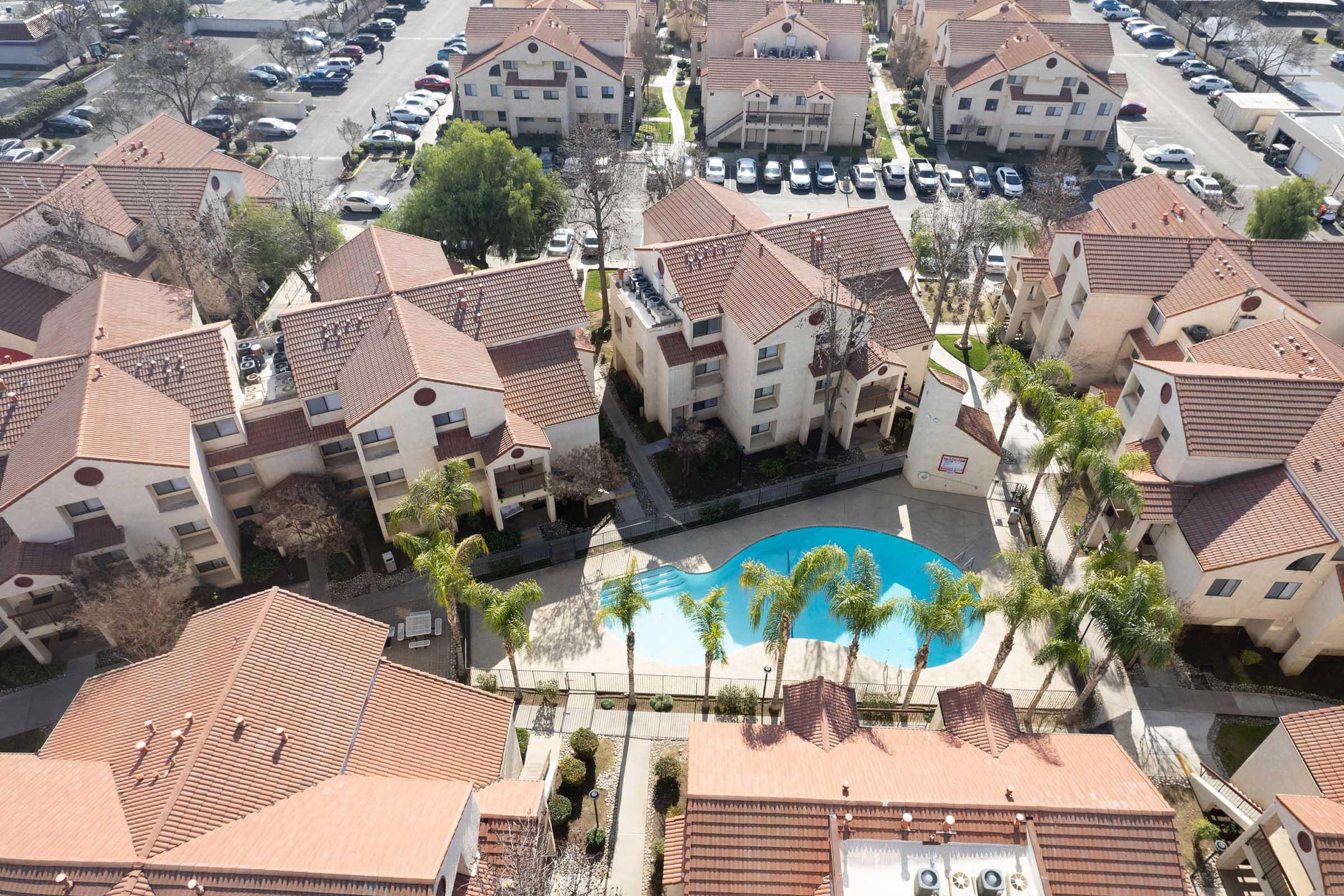 Aerial view of a residential complex featuring multiple buildings with red-tiled roofs, a central swimming pool surrounded by palm trees, and parking spaces visible in the background. The area appears well-maintained with green landscaping.