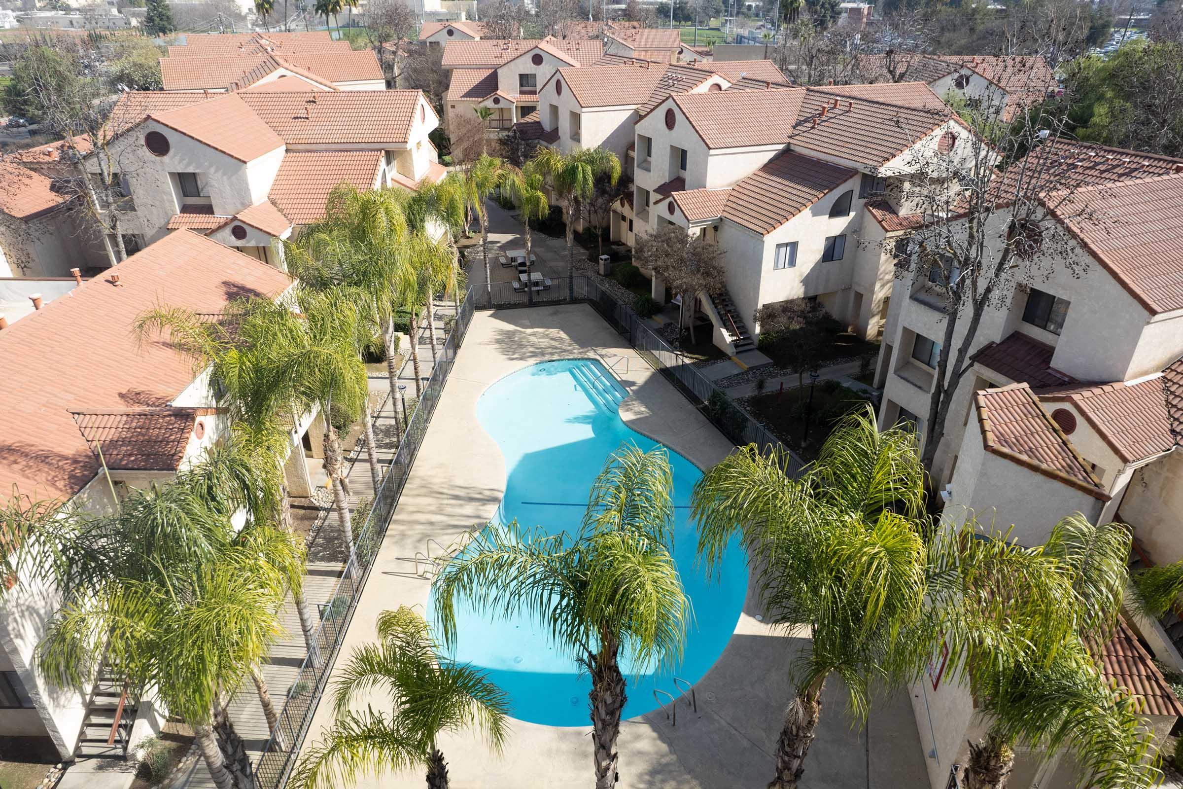 Aerial view of a residential complex featuring several buildings with red-tiled roofs. In the center, there is a swimming pool surrounded by palm trees and lounge chairs. The area is well-maintained, with green landscaping and a clear blue sky above.