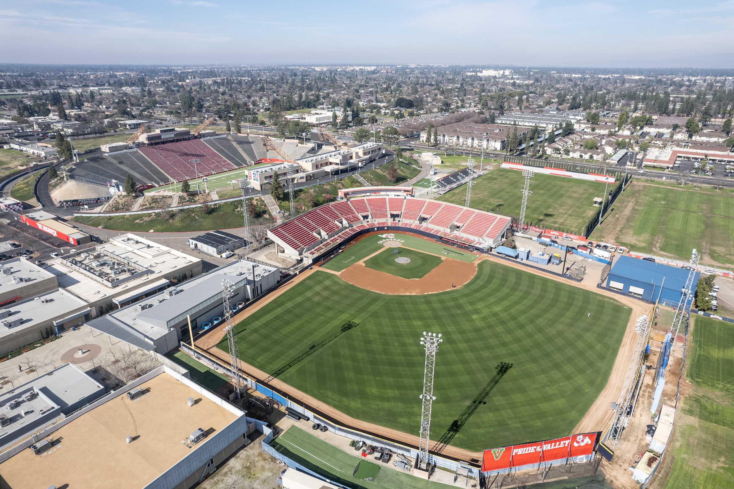 Aerial view of a baseball stadium featuring a well-maintained field, surrounded by seating areas and lights. Nearby, there are additional sports fields and urban landscape. The image shows a mix of green grass, red seating, and surrounding buildings, illustrating a sports complex with multiple facilities.
