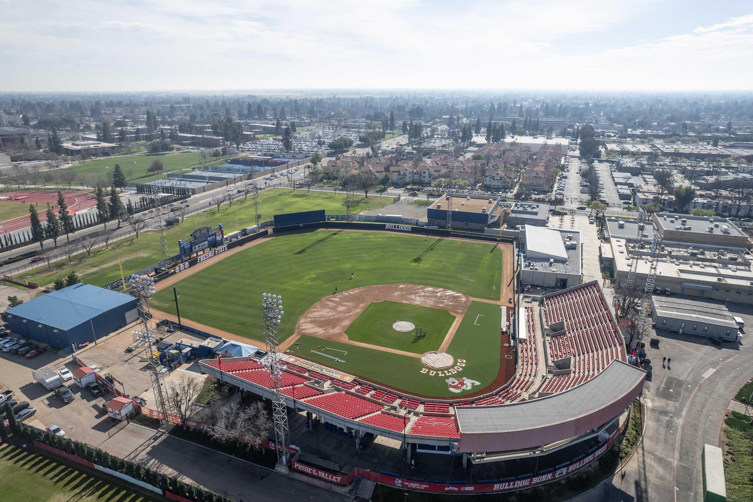 Aerial view of a baseball stadium with a well-maintained grass field, dirt infield, and a home plate. Surrounding the field are stands with red seating and a backdrop of a suburban area featuring residential buildings and streets. Clear skies above, creating a bright and sunny atmosphere.