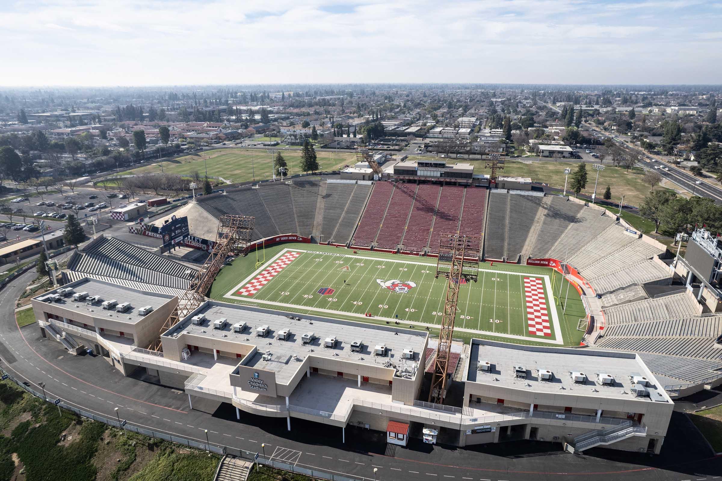 Aerial view of a large stadium featuring a well-maintained football field with red and white markings. Surrounding seating areas are visible, along with nearby sports fields and urban landscape in the background under a clear blue sky.