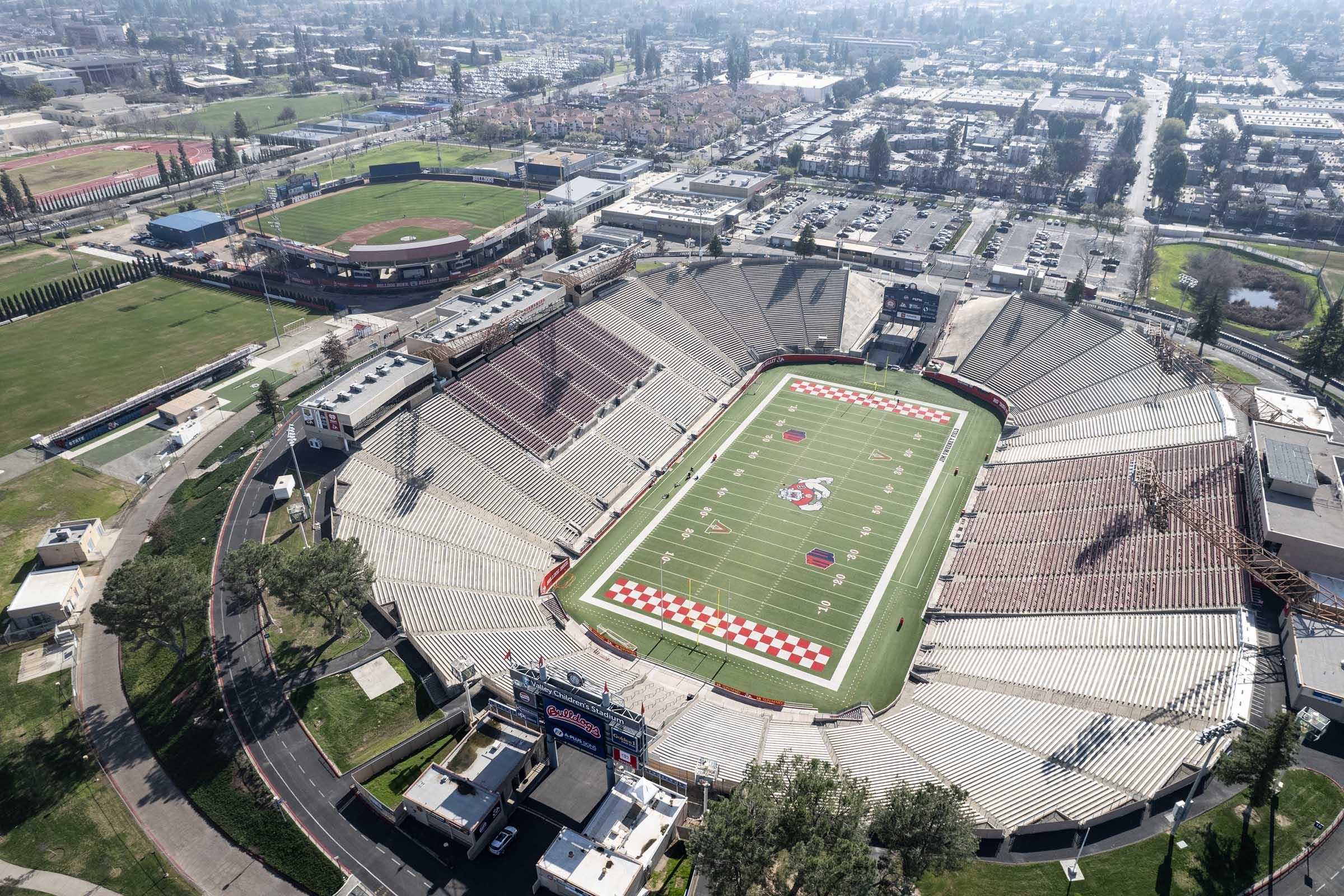 Aerial view of a large sports stadium with a football field, surrounded by seating areas and green fields. In the background, there are residential buildings and a baseball field visible. The stadium features a distinctive field design with red and white patterns.